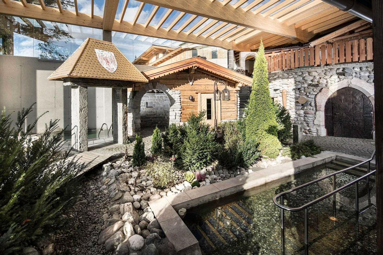 A rustic courtyard with stones, plants, and a small pond. In the background, wooden structures and a traditional roof are visible.