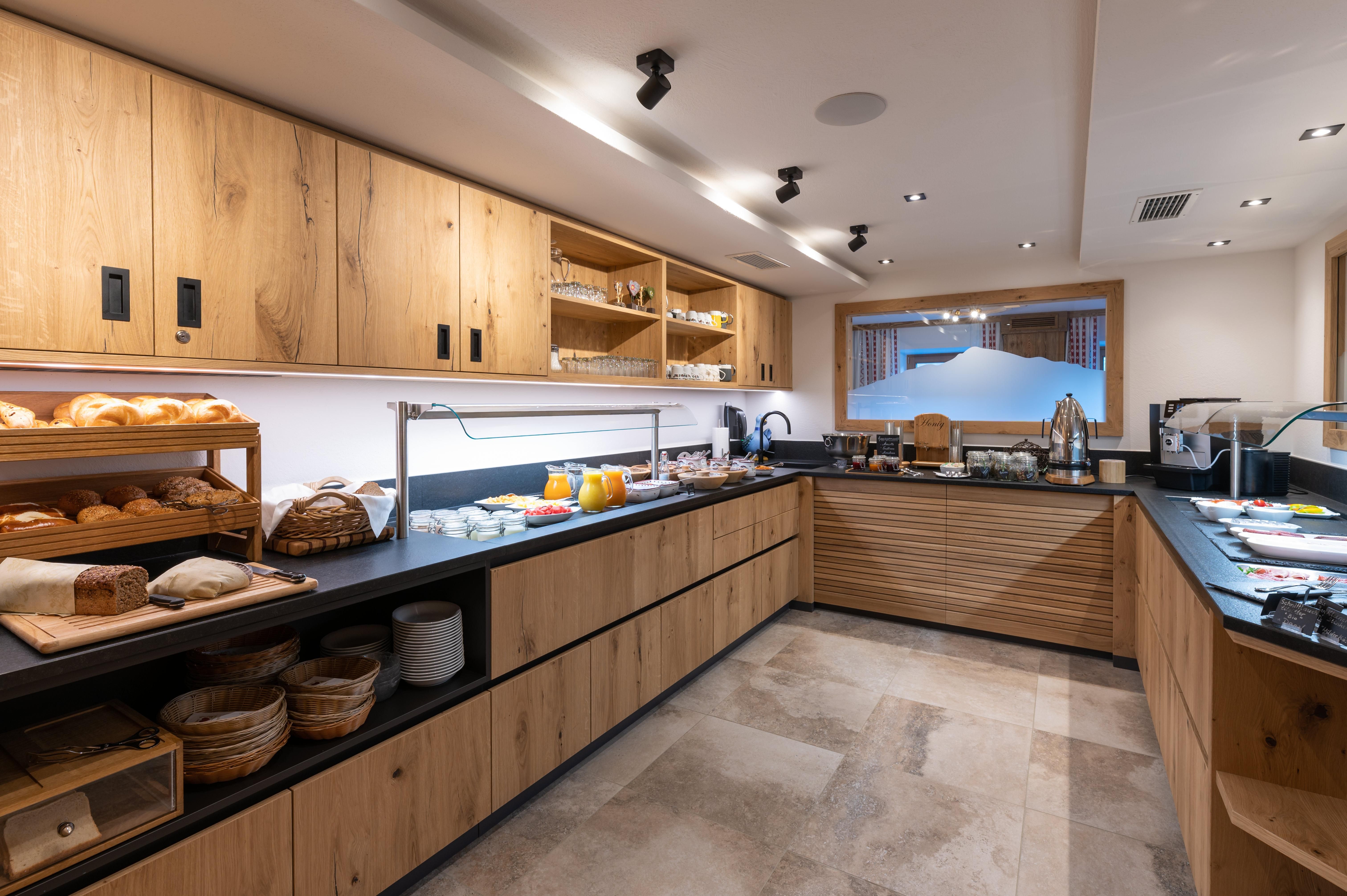 A modern kitchen with wooden cabinets and an extensive breakfast buffet selection. Fresh bread, fruits, beverages, and dishes are visible on the countertop.