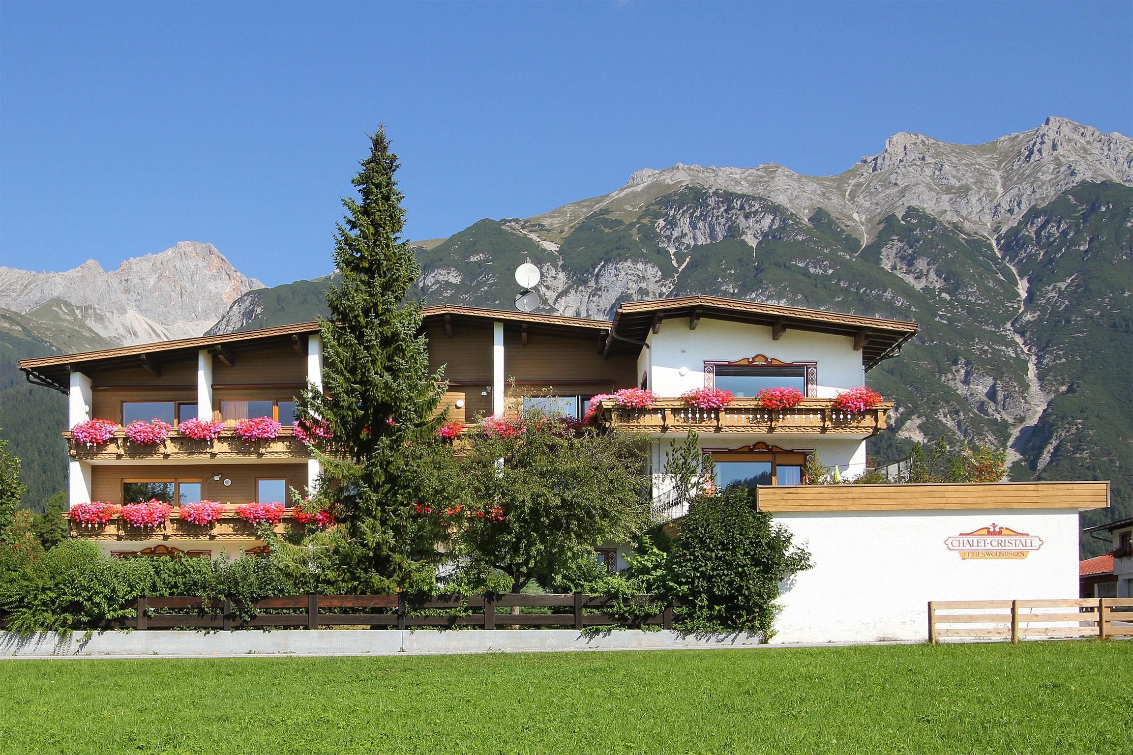 A cozy building surrounded by green meadows and majestic mountains. The balconies are adorned with colorful flowers, creating an inviting atmosphere.