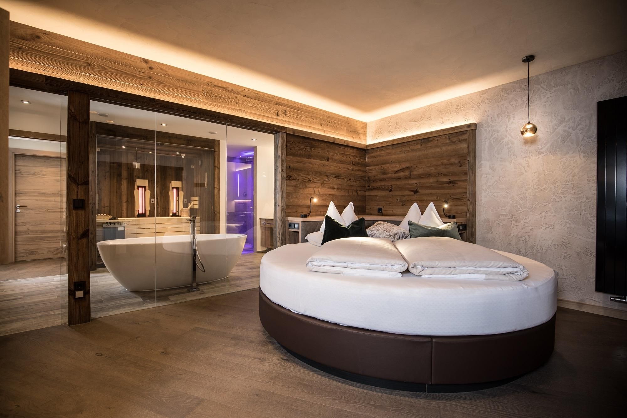 A modern hotel room with a round bed and wood paneling. In the background, there is an elegant bathroom with a freestanding bathtub.