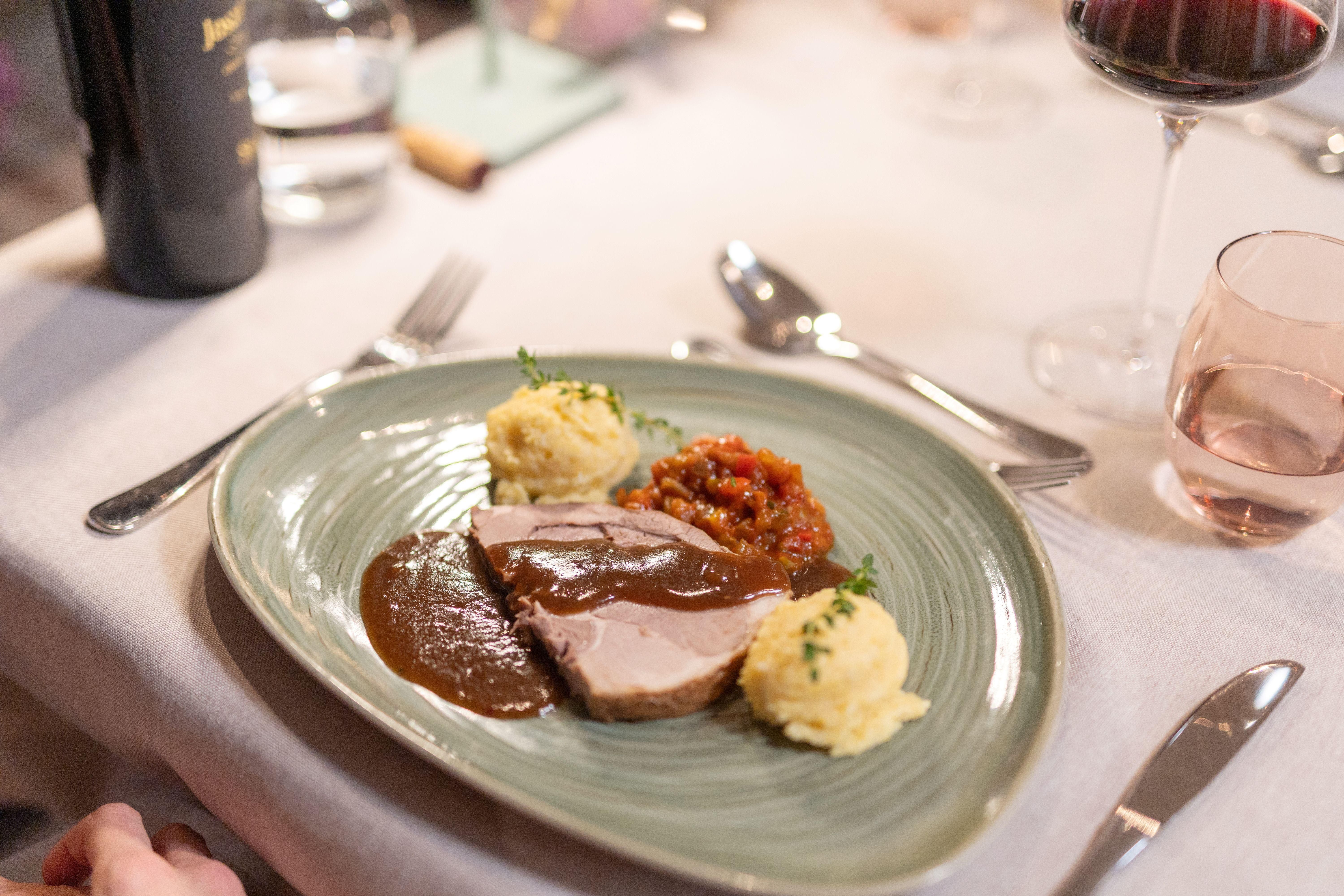 A delicious plate of tender meat in a brown sauce, served with mashed potatoes and a tomato sauce. The table is elegantly set with cutlery and glasses.