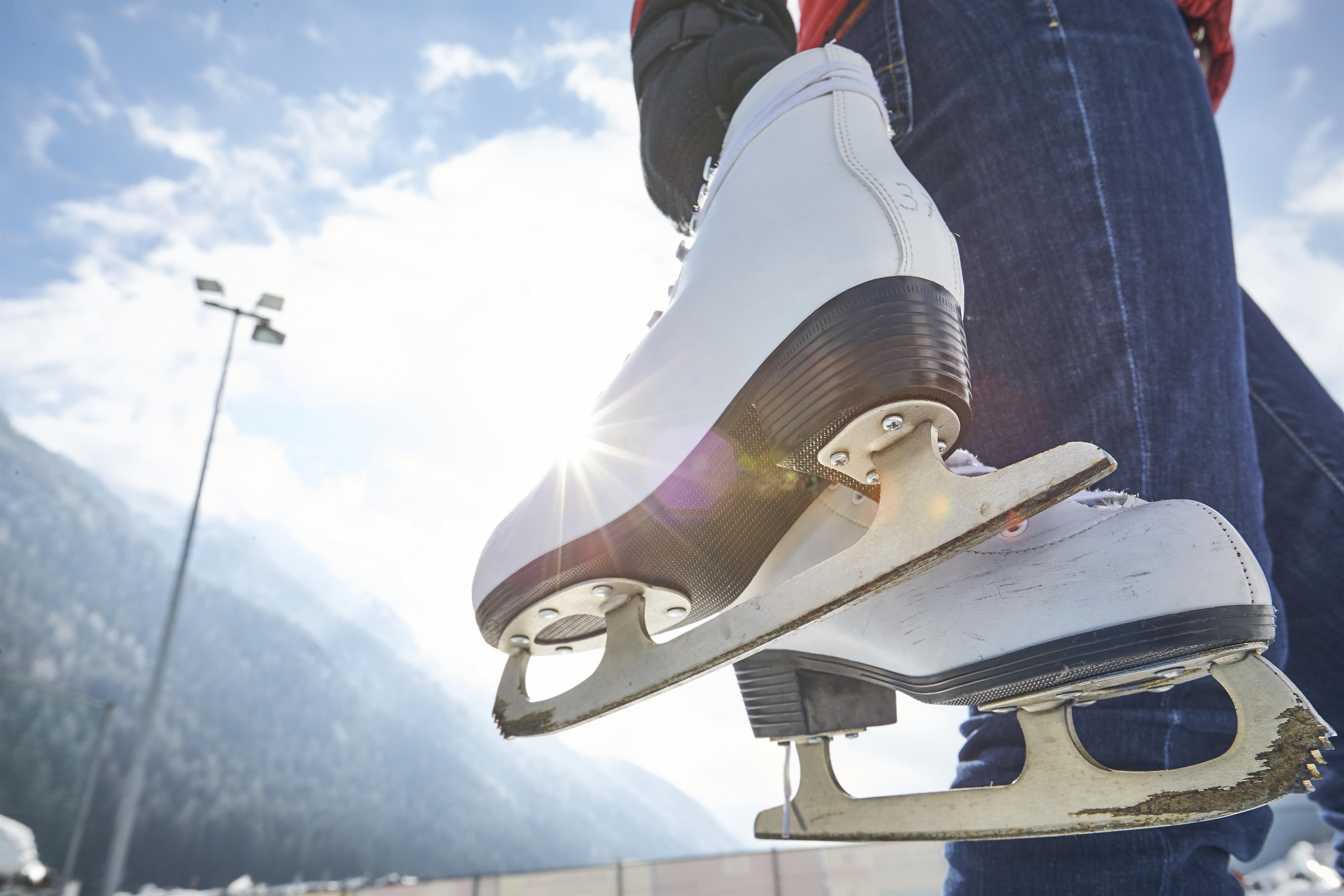 A ice skater is holding a white skate in their hand. In the background, there are mountains and a clear sky.