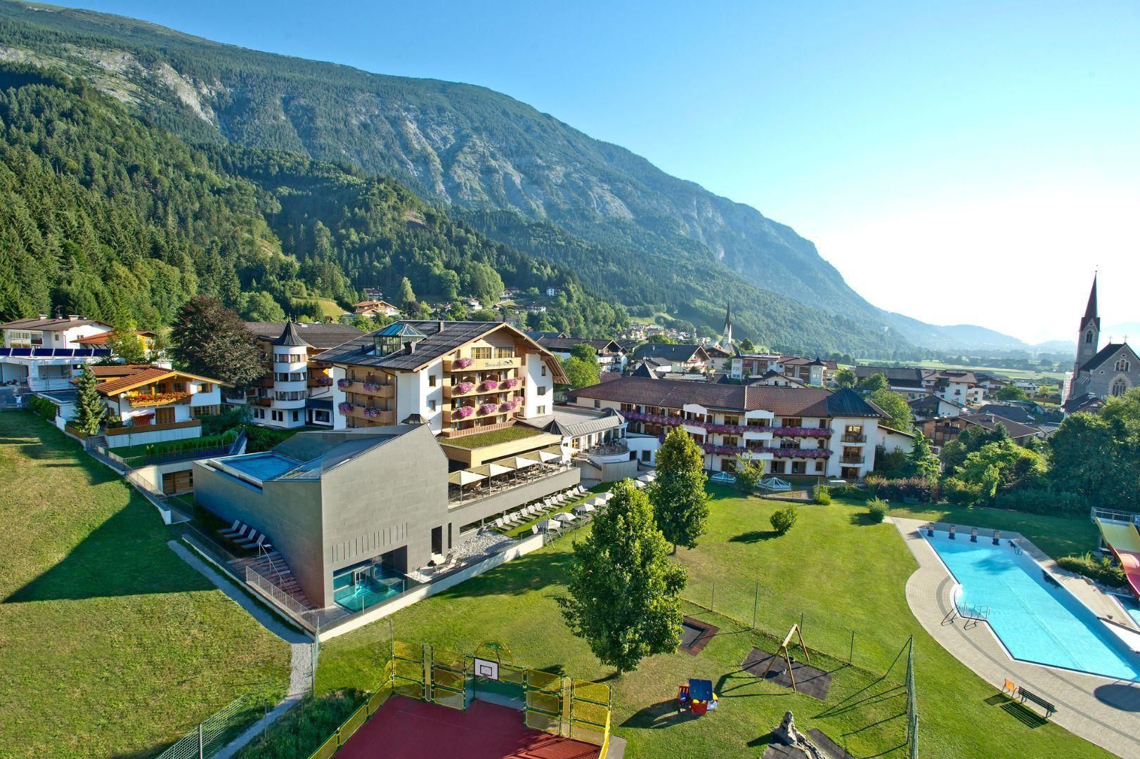 A picturesque mountain village with modern hotels and a green environment. In the foreground, a swimming pool is visible, surrounded by meadows and trees.