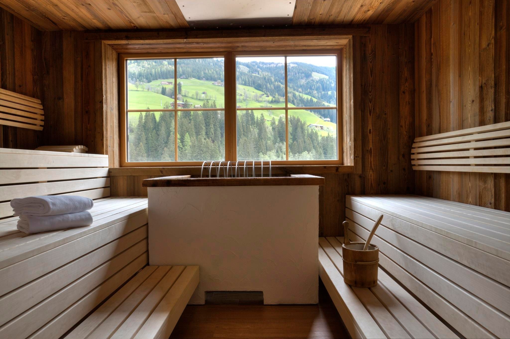 A bright sauna room with wooden paneling and a large window front. The view extends over green hills and forests.