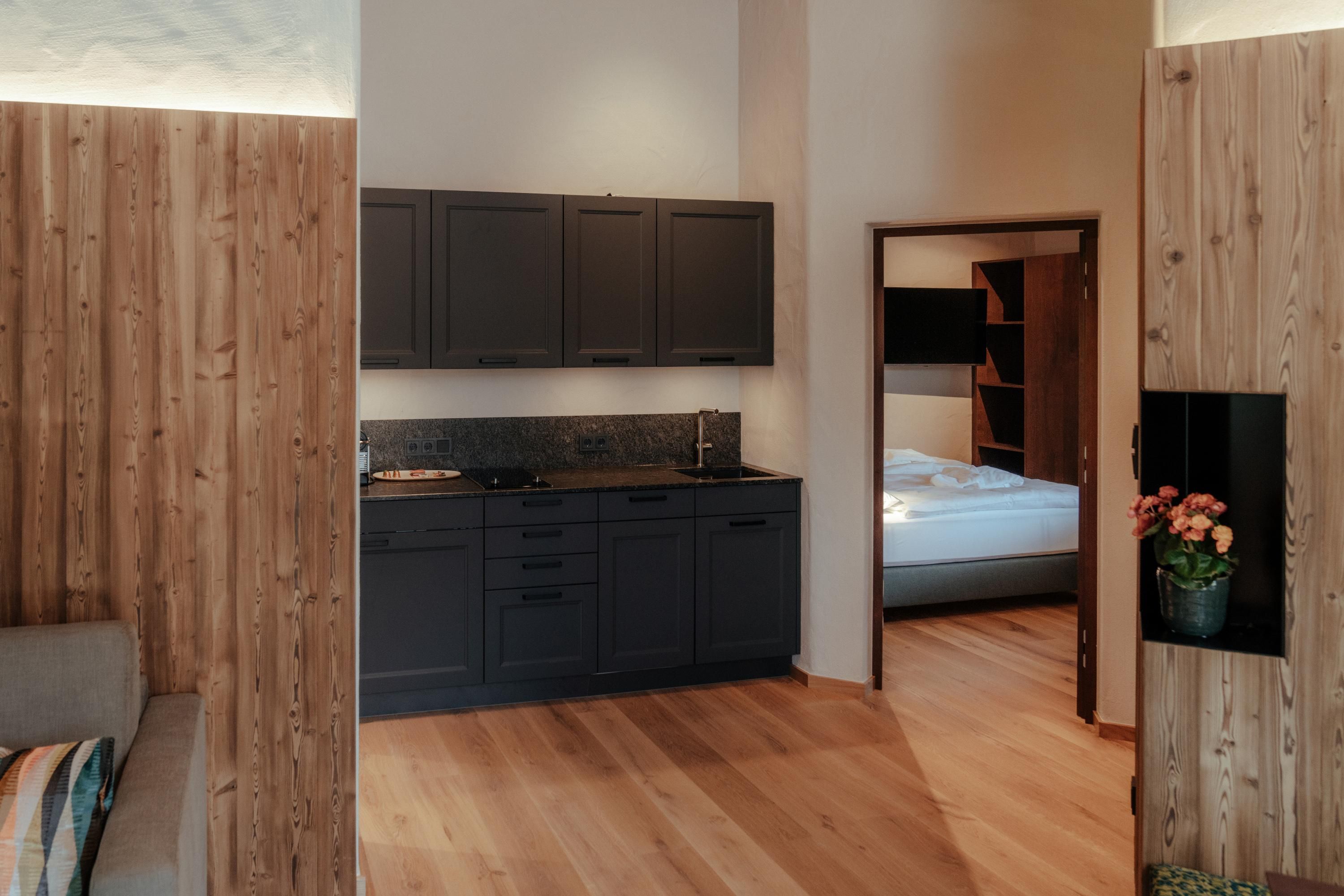 A modern kitchen with dark cabinets and wood paneling. In the background, a bedroom with a bed is visible.