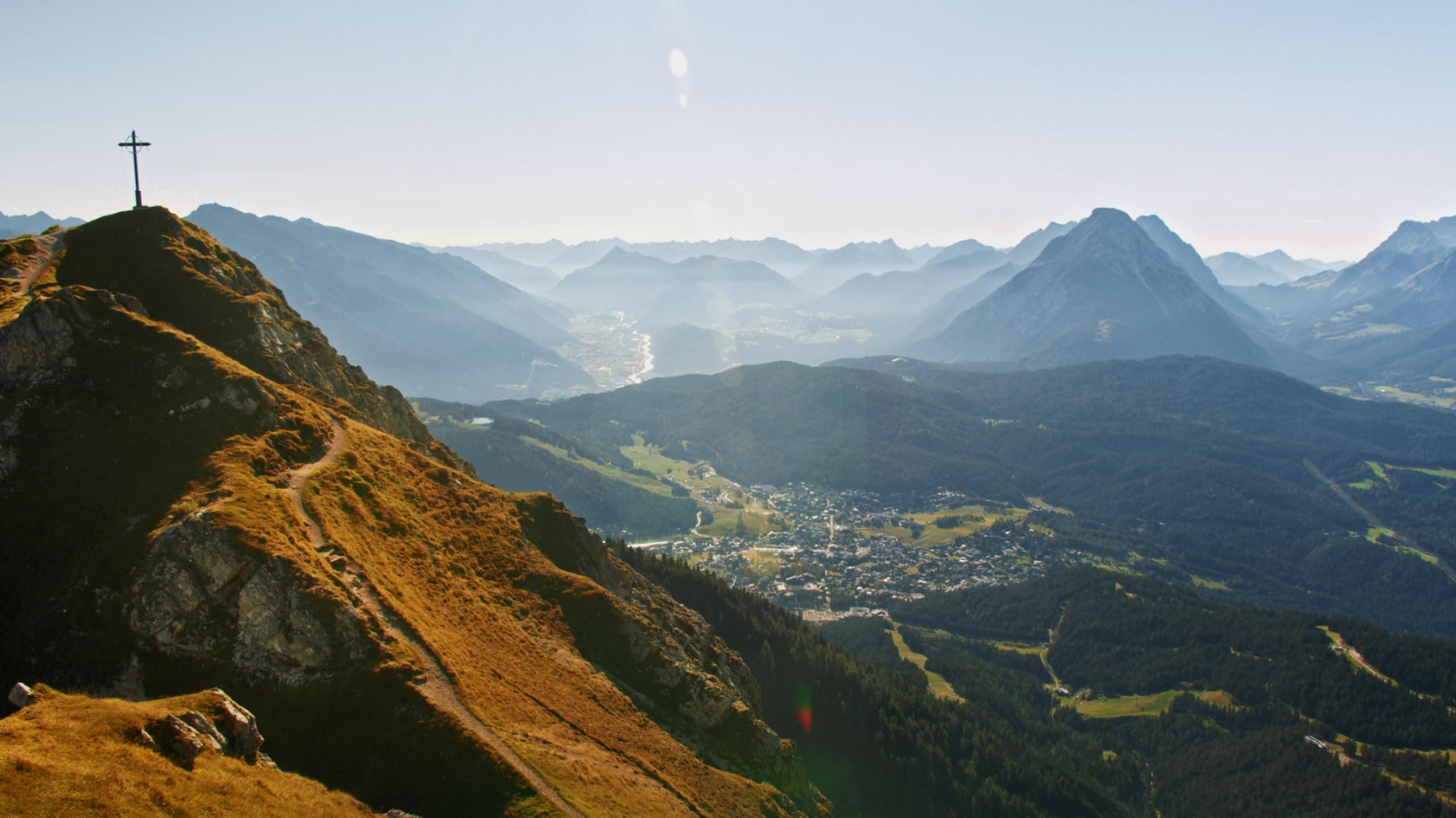 A picturesque mountain landscape with high peaks and a clear sky. In the foreground, there is a hill with a cross and a quiet valley town.