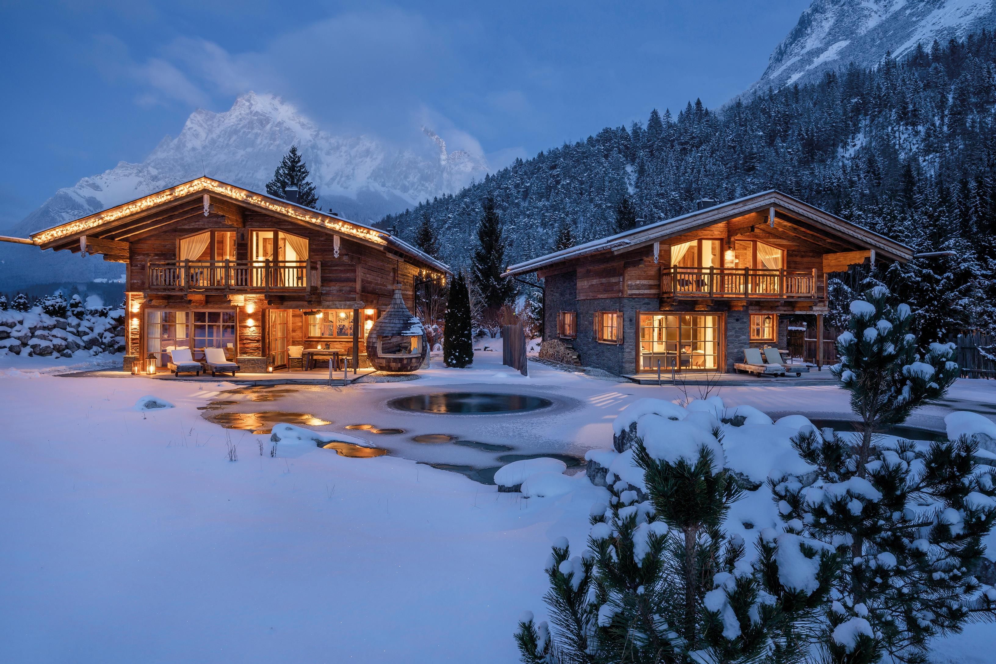 Two cozy cabins in the snowy winter forest, illuminated by warm light. In the background, majestic mountains rise.
