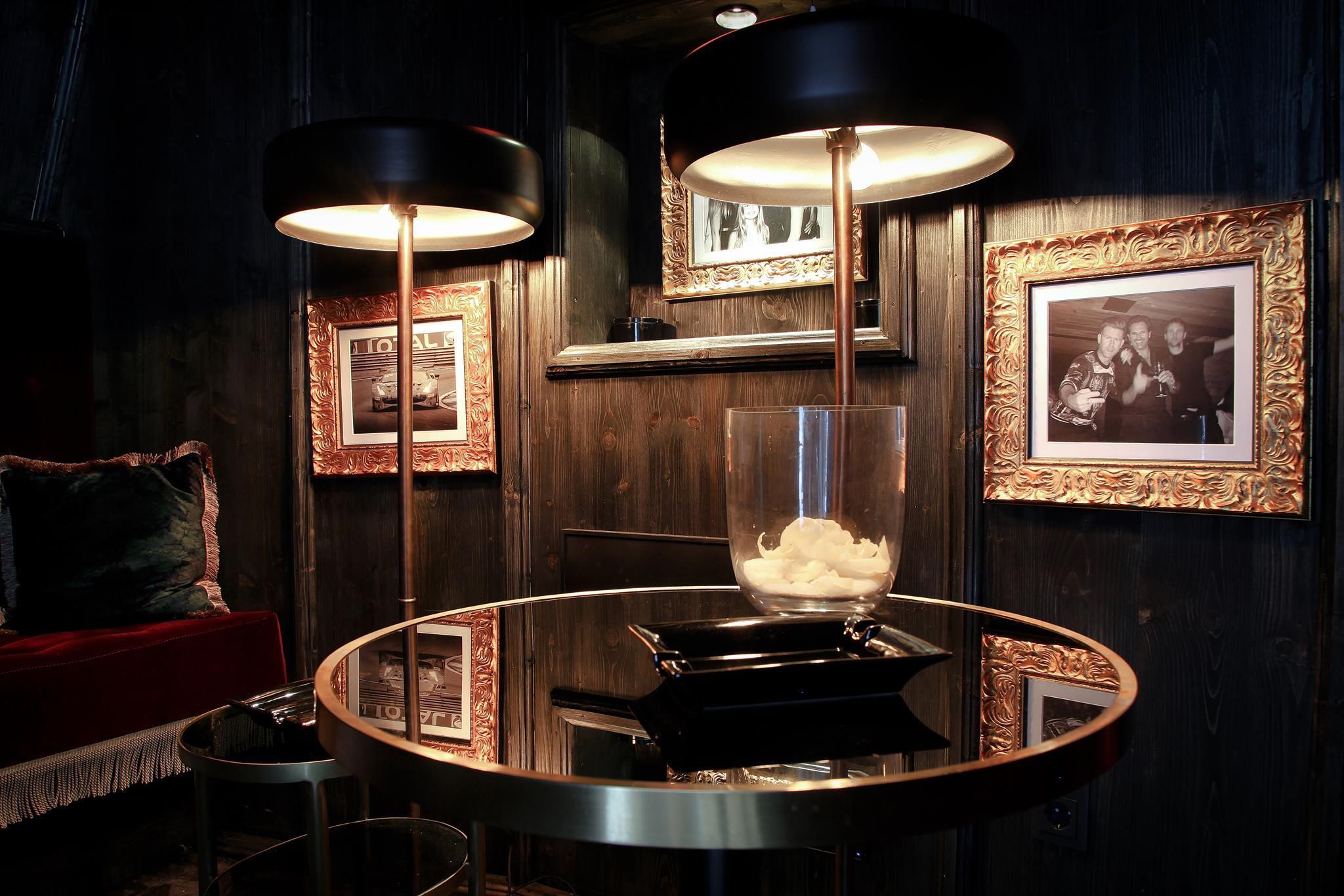 A stylish room with two modern lamps and a round table. The walls are adorned with elegant, gold picture frames.