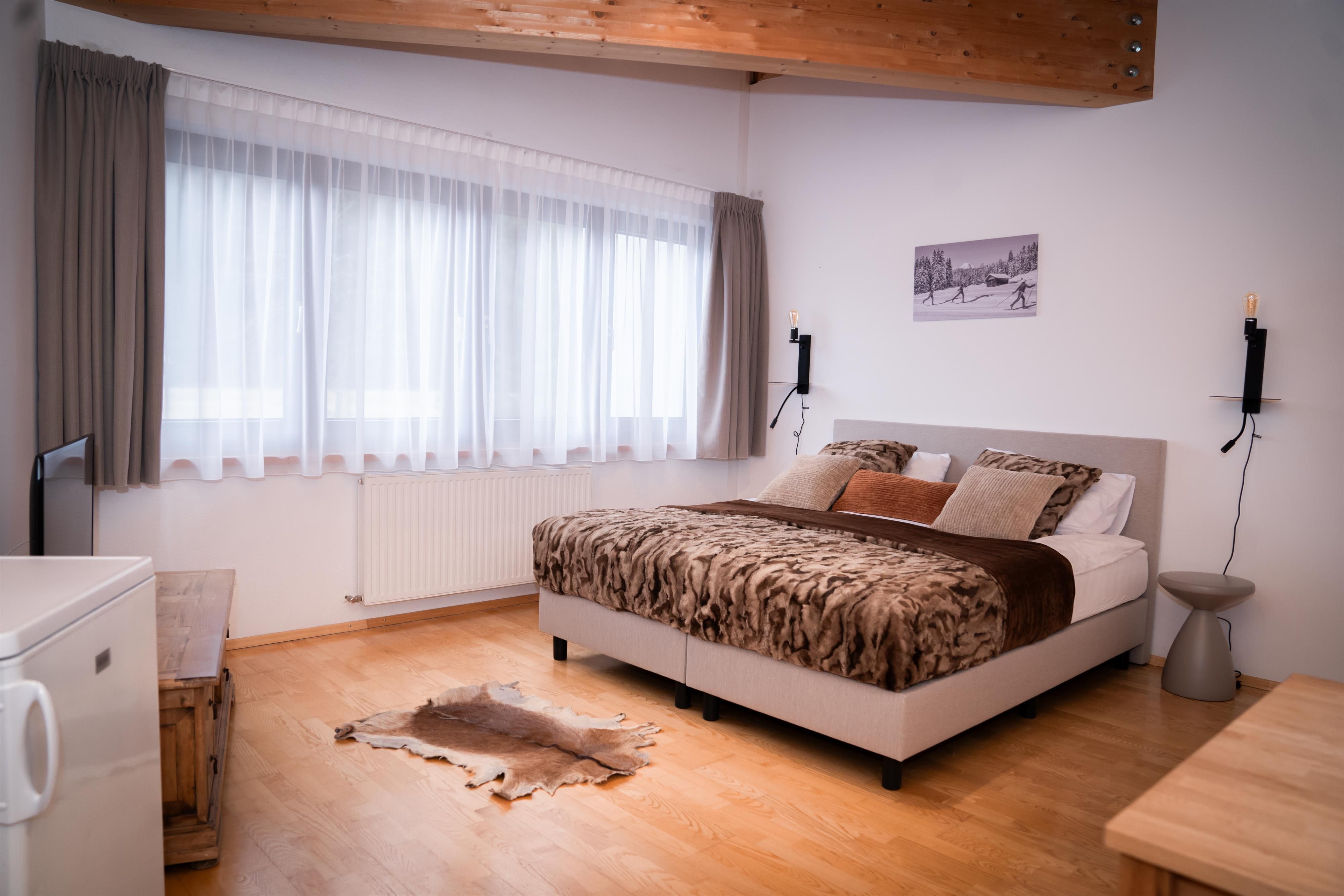 A cozy bedroom with a large bed and light wooden floor. There are large windows with curtains and a modern decor.