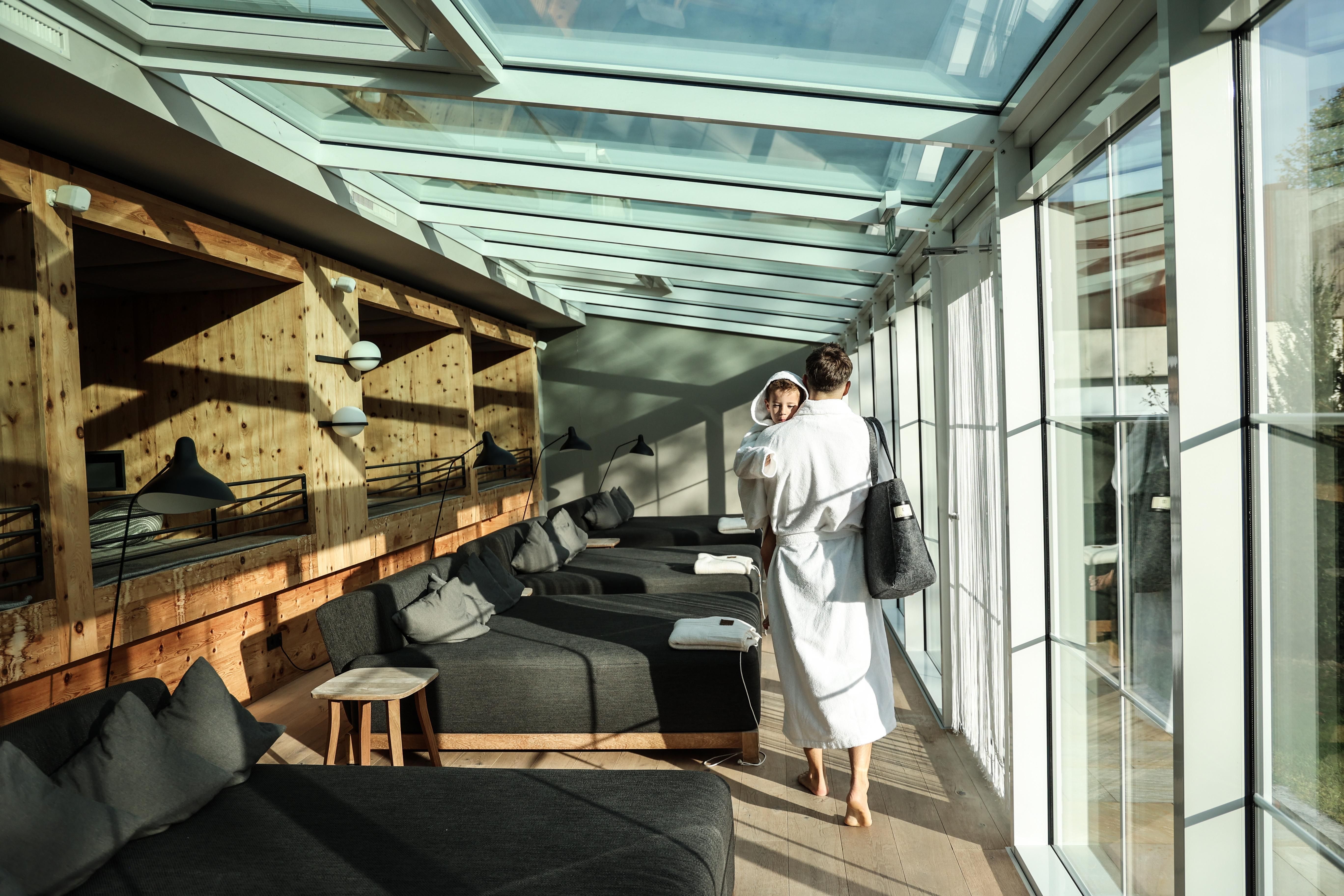 A bright, modern relaxation room with large windows and cozy loungers. A person in a bathrobe is carrying a towel and walking through the room.