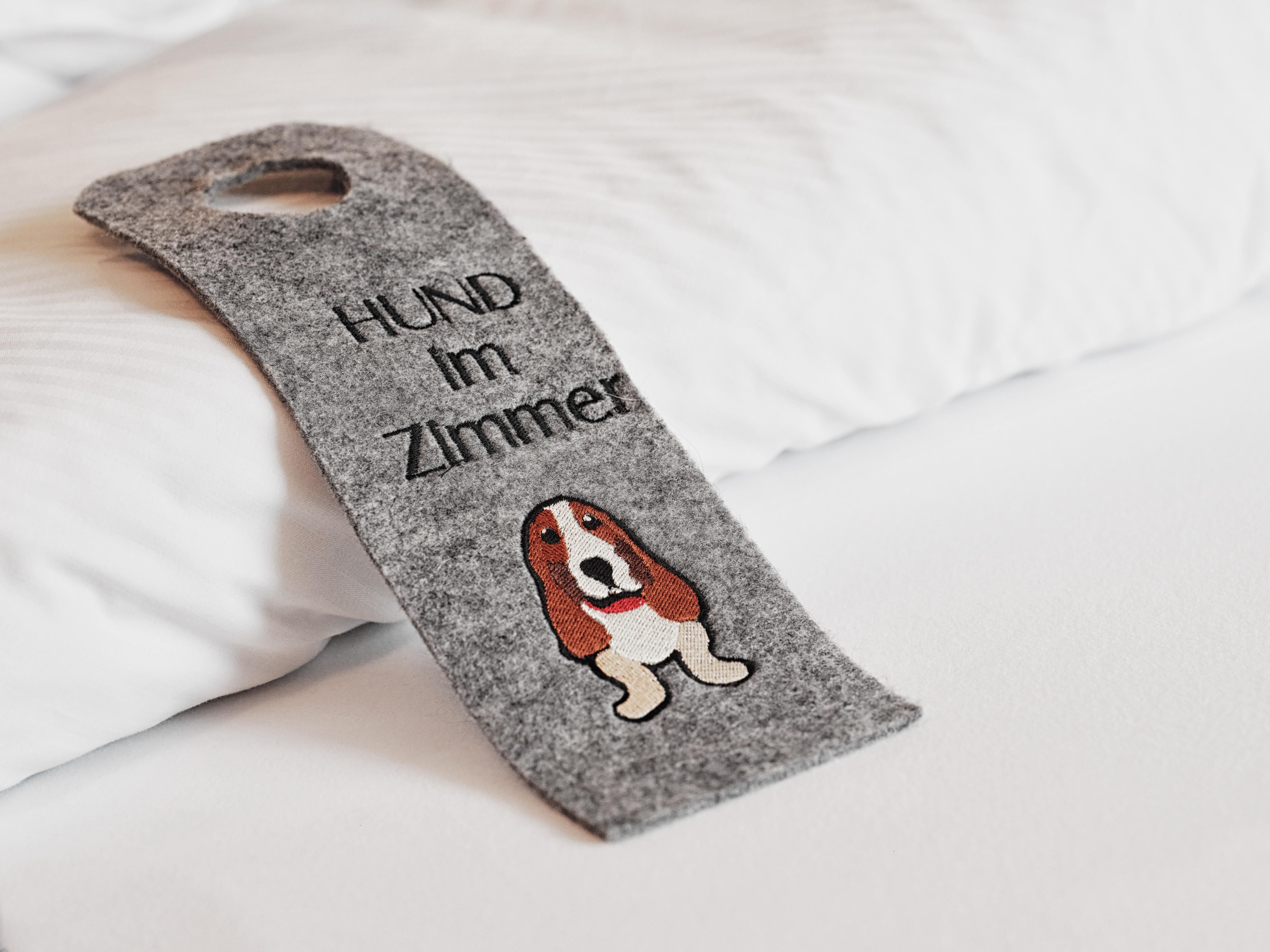 A gray sign with the inscription "DOG in the room" and an image of a dog icon. It is lying on a bed with a white pillow.