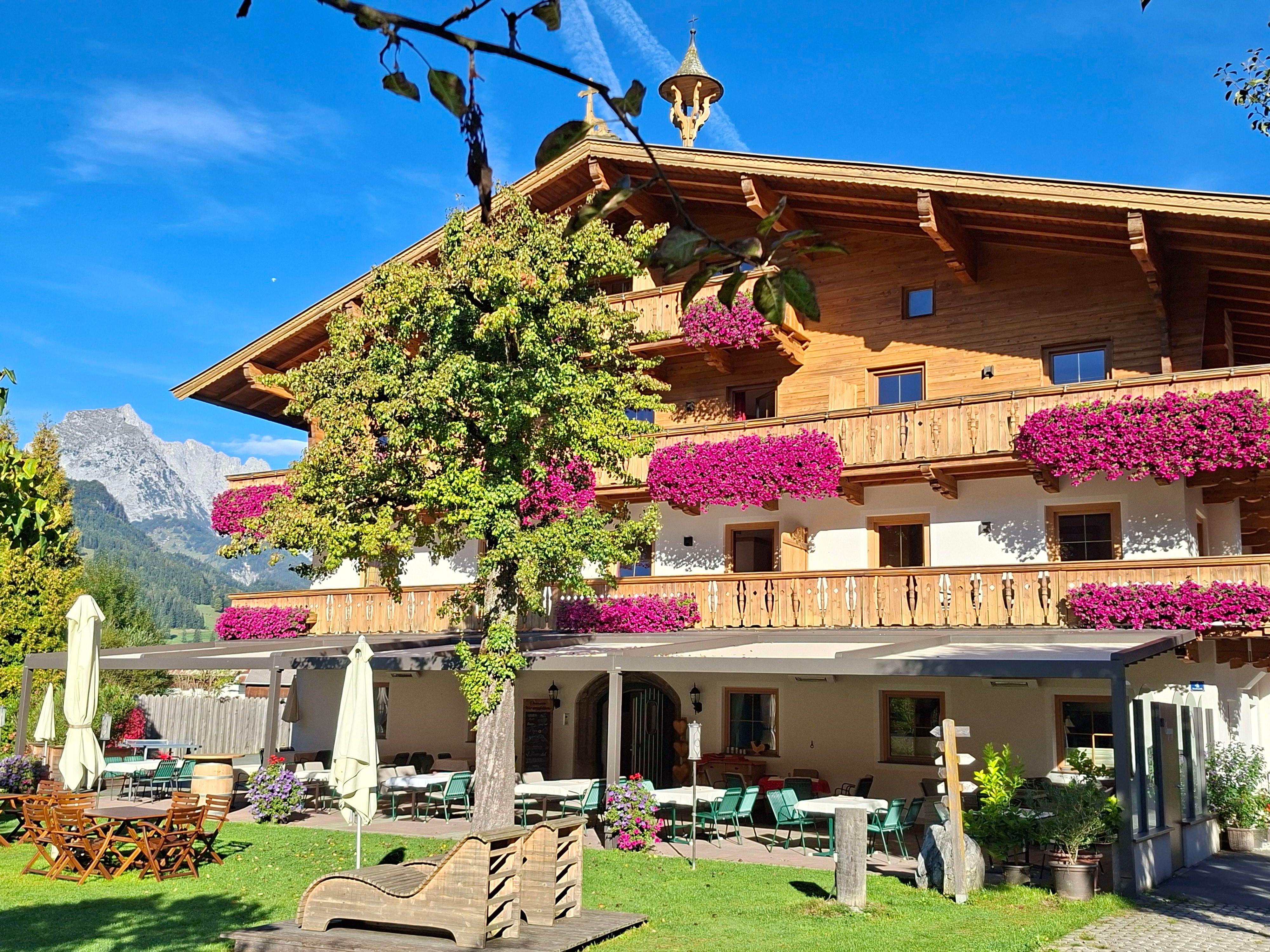 A charming wooden building with colorful flowers on the balconies. The surroundings are green and offer a view of the mountains in the background.
