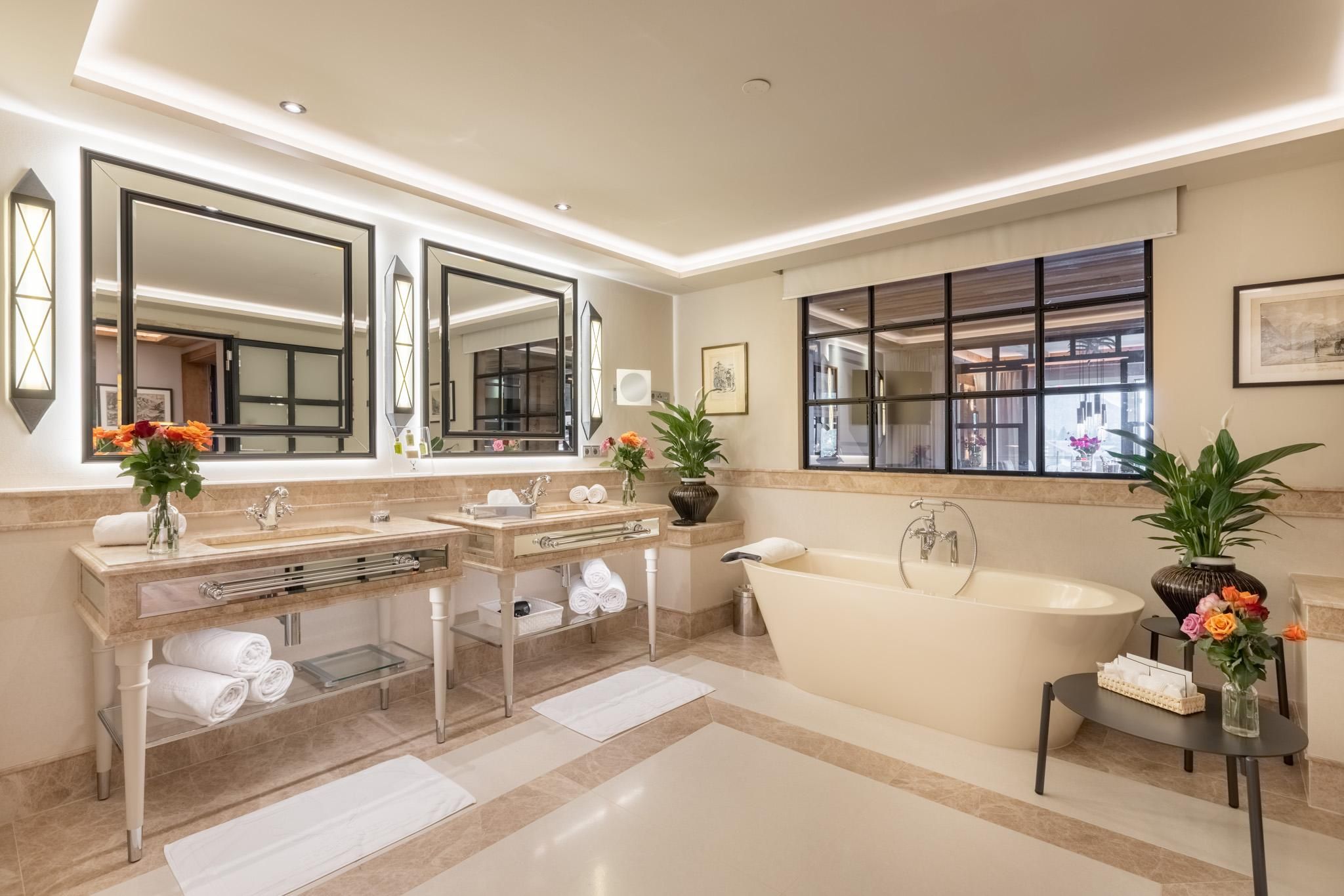 A modern bathroom with an elegant bathtub and two sinks. It also features fresh flowers and large mirrors that provide light and space.
