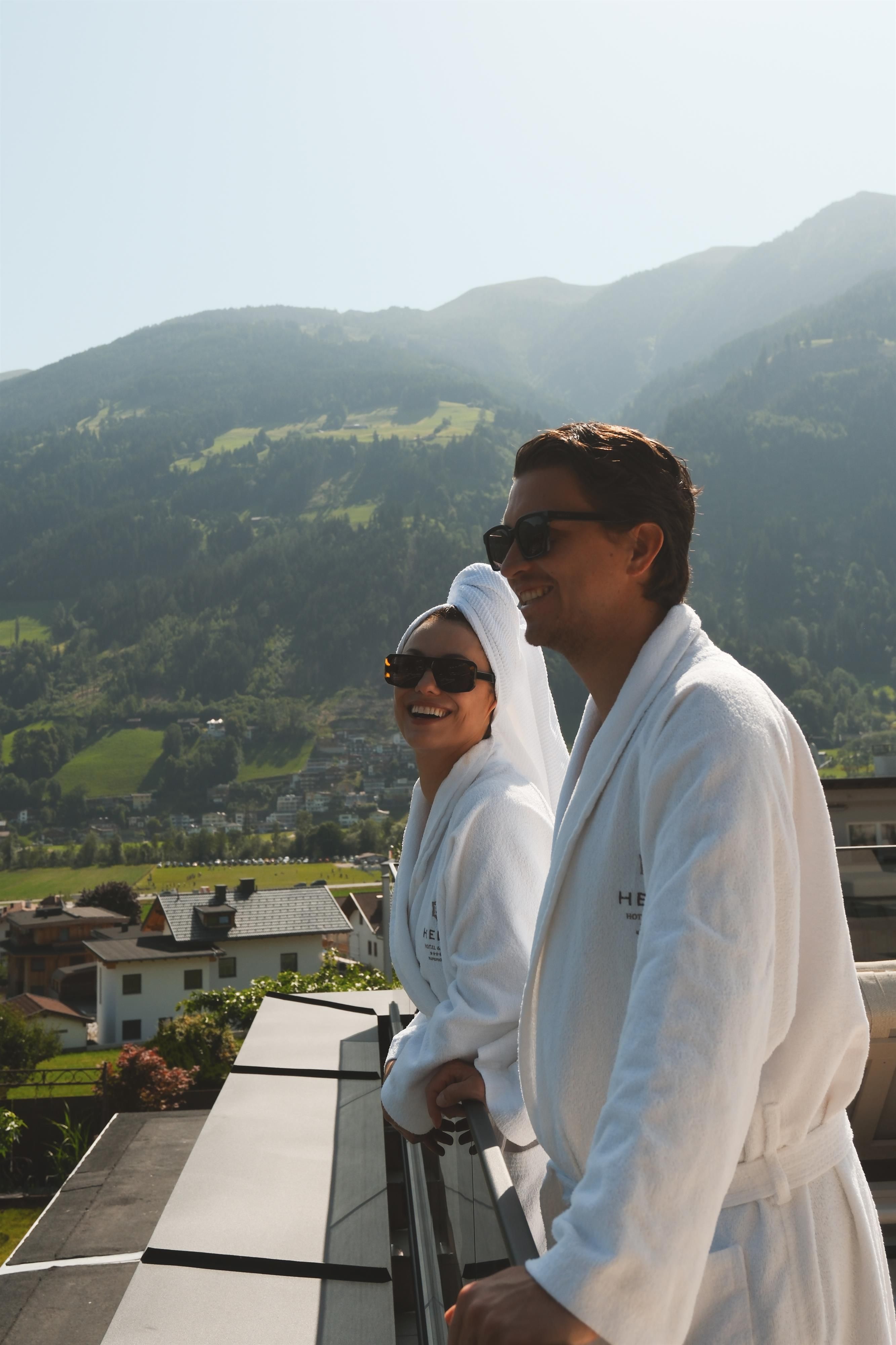 A couple in bathrobes stands on a balcony and looks out at the picturesque mountain landscape. The sky is clear and the sun is shining.
