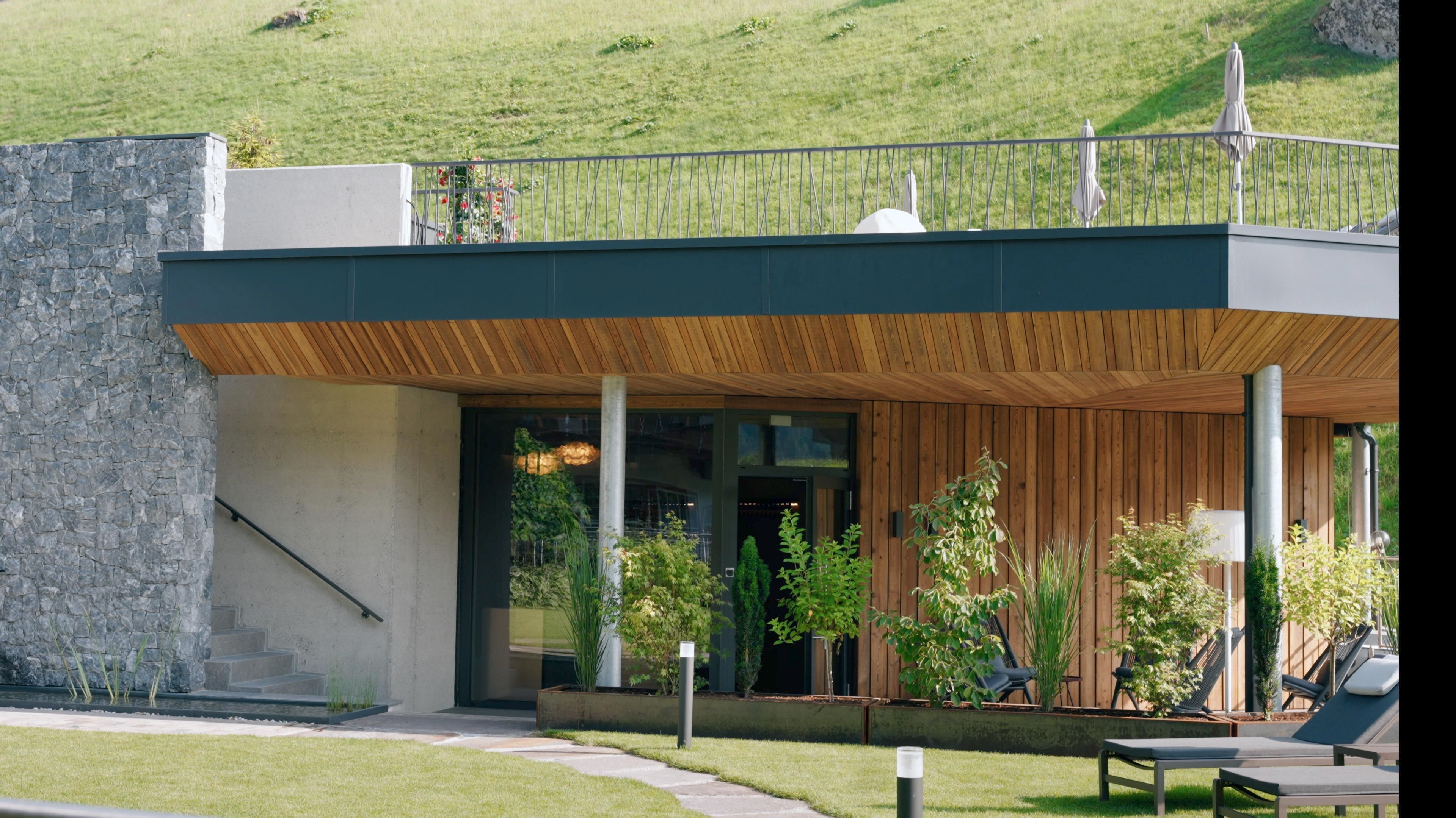A modern building with wood and stone elements in a green environment. The terrace offers a view of the surrounding landscape.