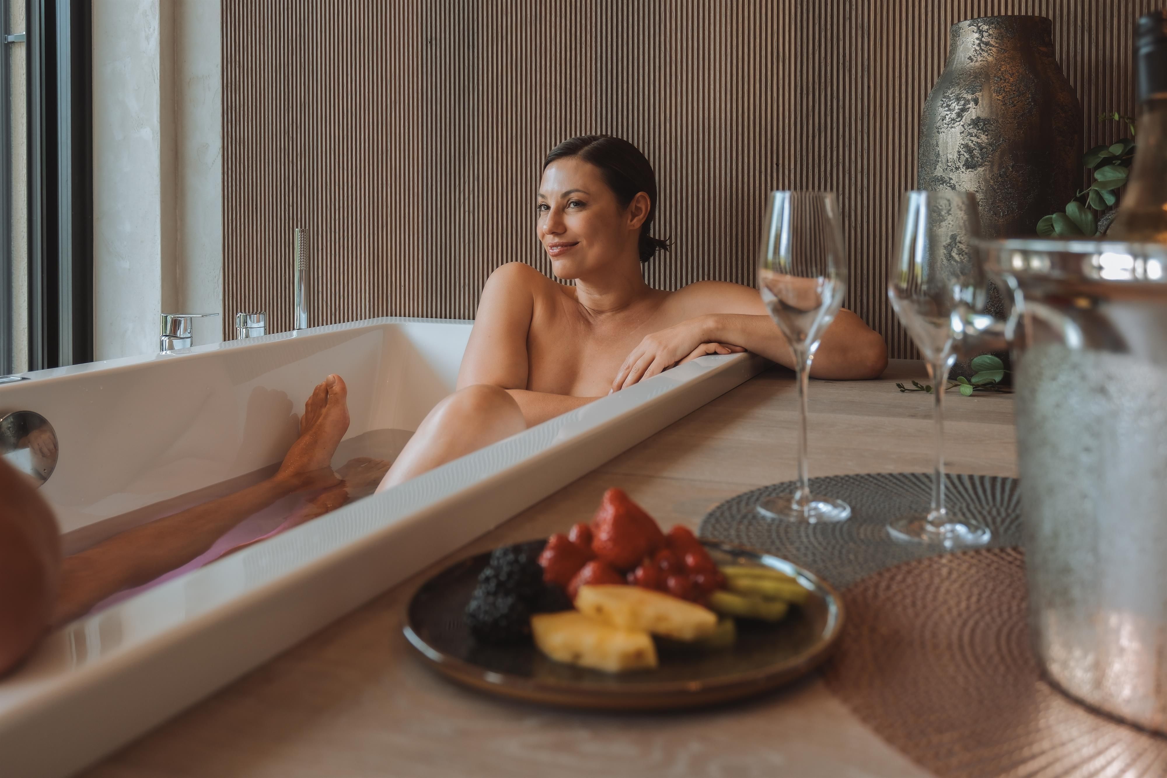 A woman relaxes in a bathtub and enjoys her time off. On the table is a platter of fresh fruit and glasses.