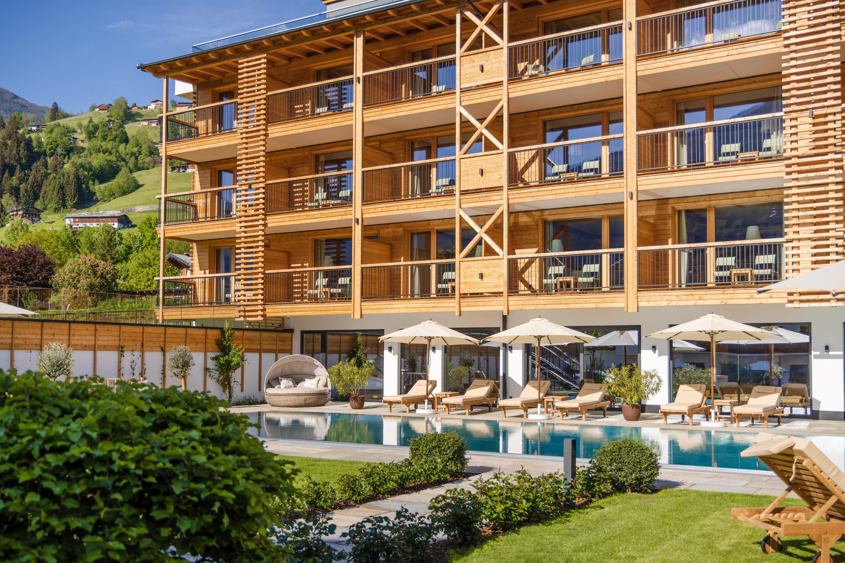 A modern hotel with a wooden facade and large windows. In the foreground, there is a beautiful pool with loungers and sun umbrellas.