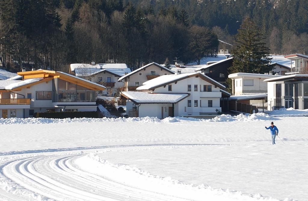A winter landscape with snow-covered houses and a clear blue sky. A person is walking on the snow near the shore.