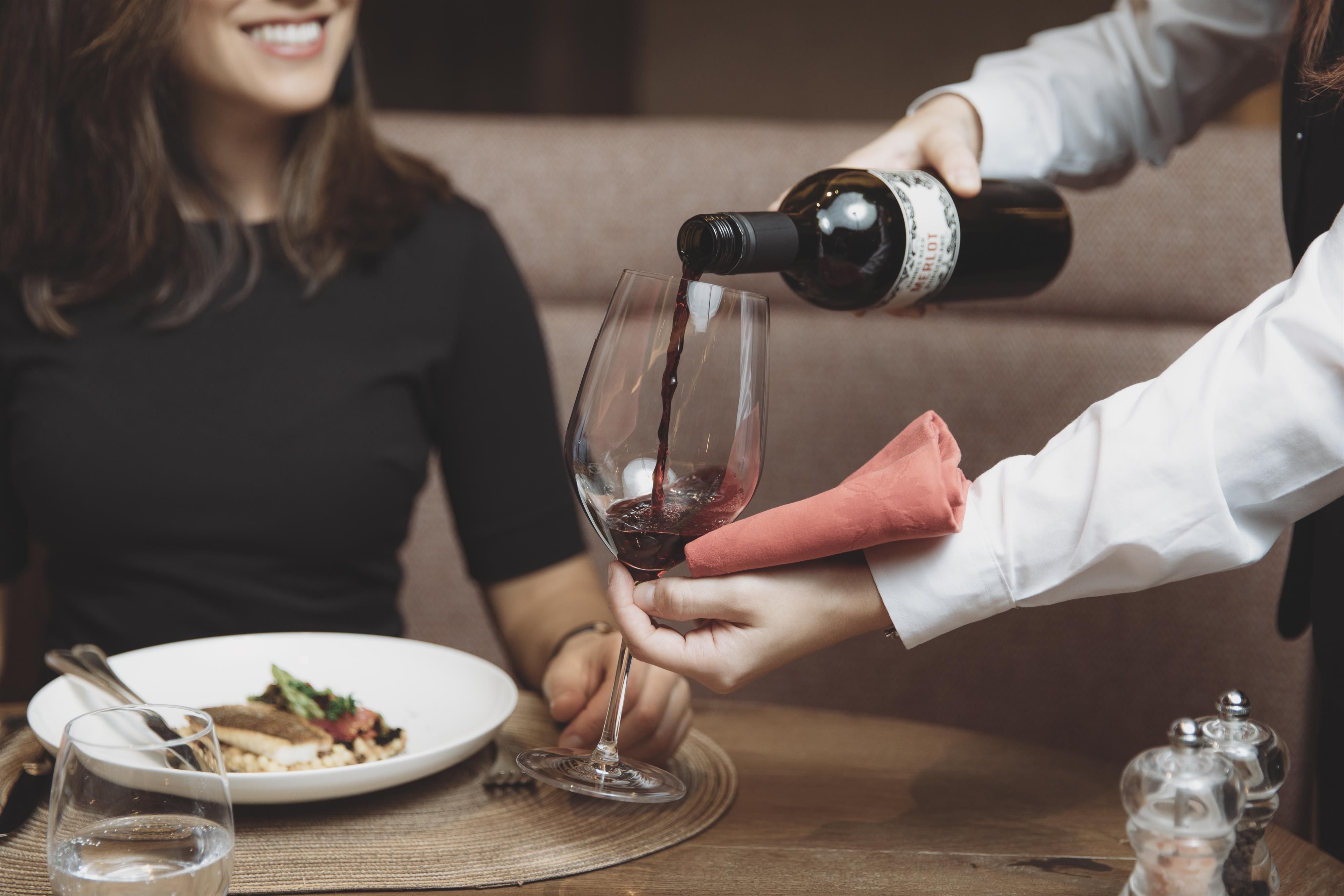A waiter pours red wine into a glass while a person at the table smiles. In the background, a plate with a dish can be seen.