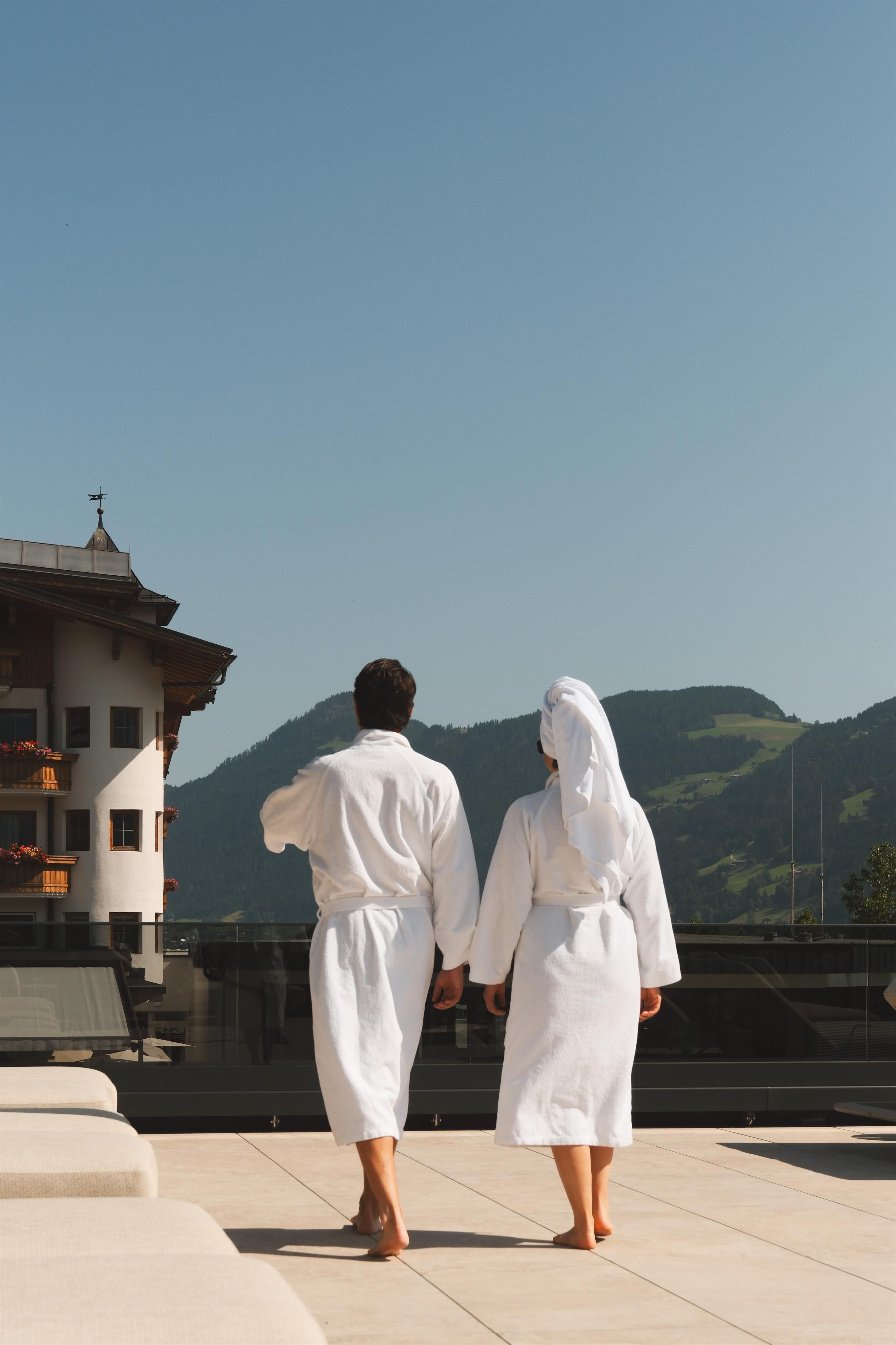 A couple in white bathrobes is walking on a terrace. In the background, green mountains and a clear sky can be seen.