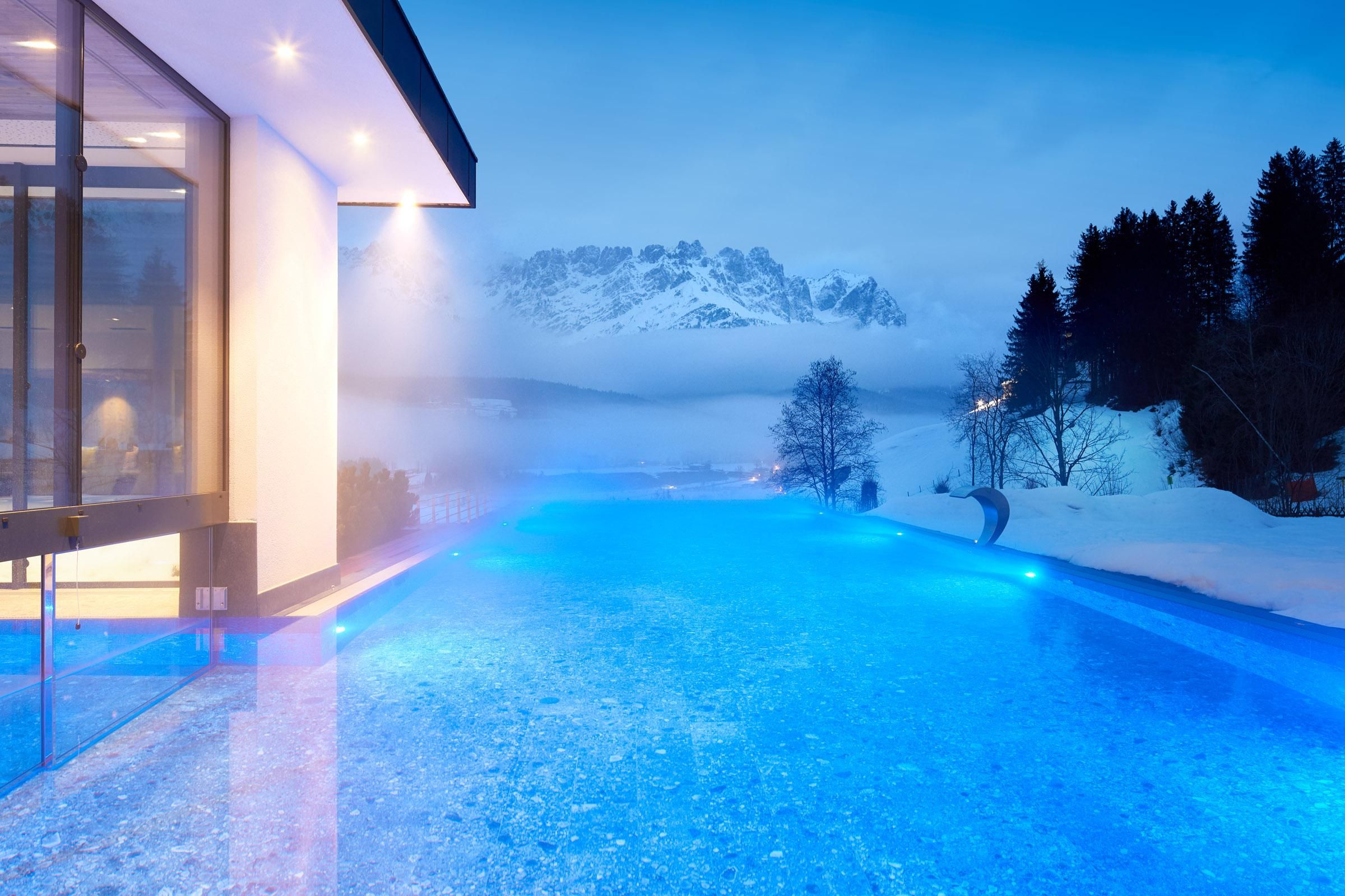 A modern house with a blue infinity pool and snow-covered mountains in the background. The landscape is calm and wintry, with a blurred view of the mountains.