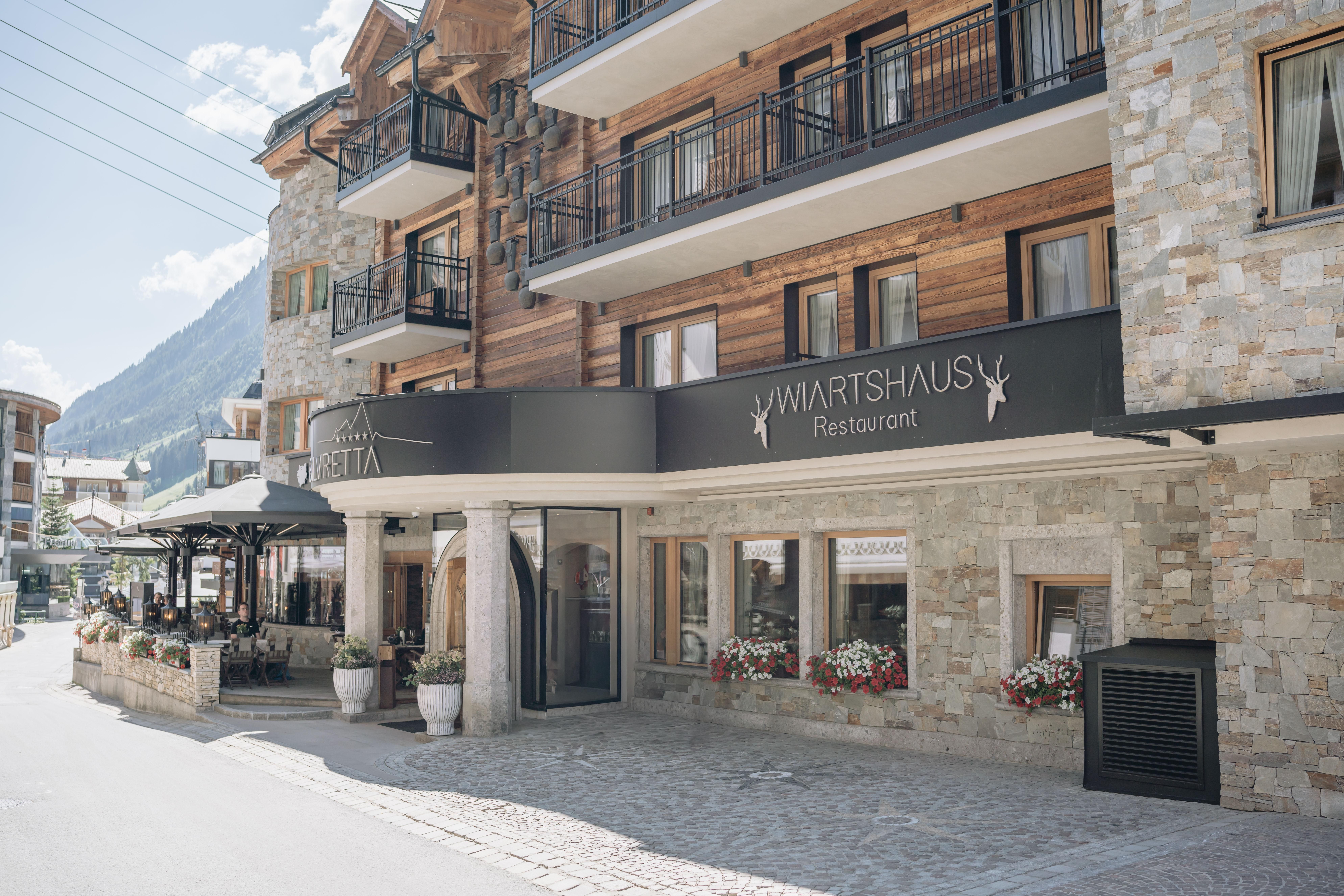 A modern restaurant with an inviting facade made of wood and stone. Outside, there are tables with flowers, and the surroundings are surrounded by mountains and a blue sky.