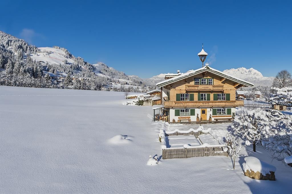 A picturesque house in winter, surrounded by a snow-covered landscape. In the background, the mountains rise under a clear blue sky.