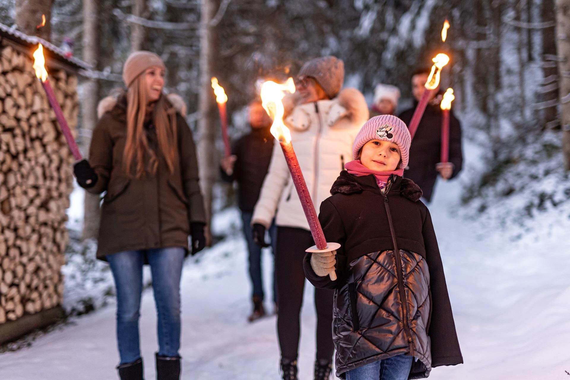 A group of people is walking through the snow while carrying torches. In the foreground stands a girl with a hat and a smile.