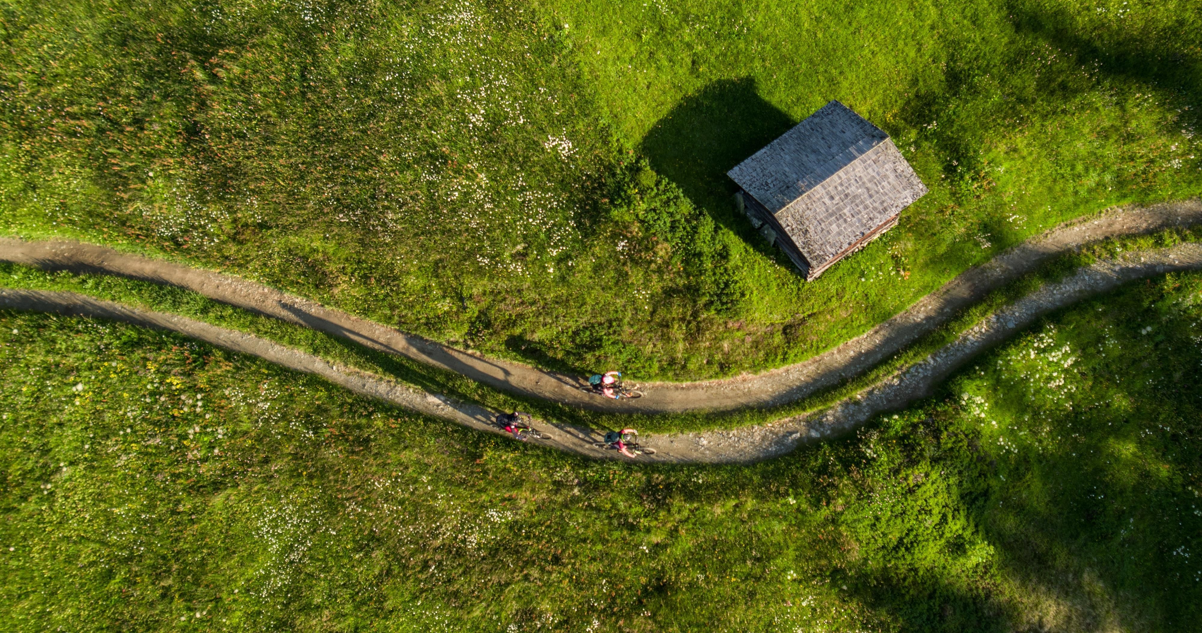 A picturesque landscape with green meadows and a small wooden house. Cyclists pass by on a winding path.