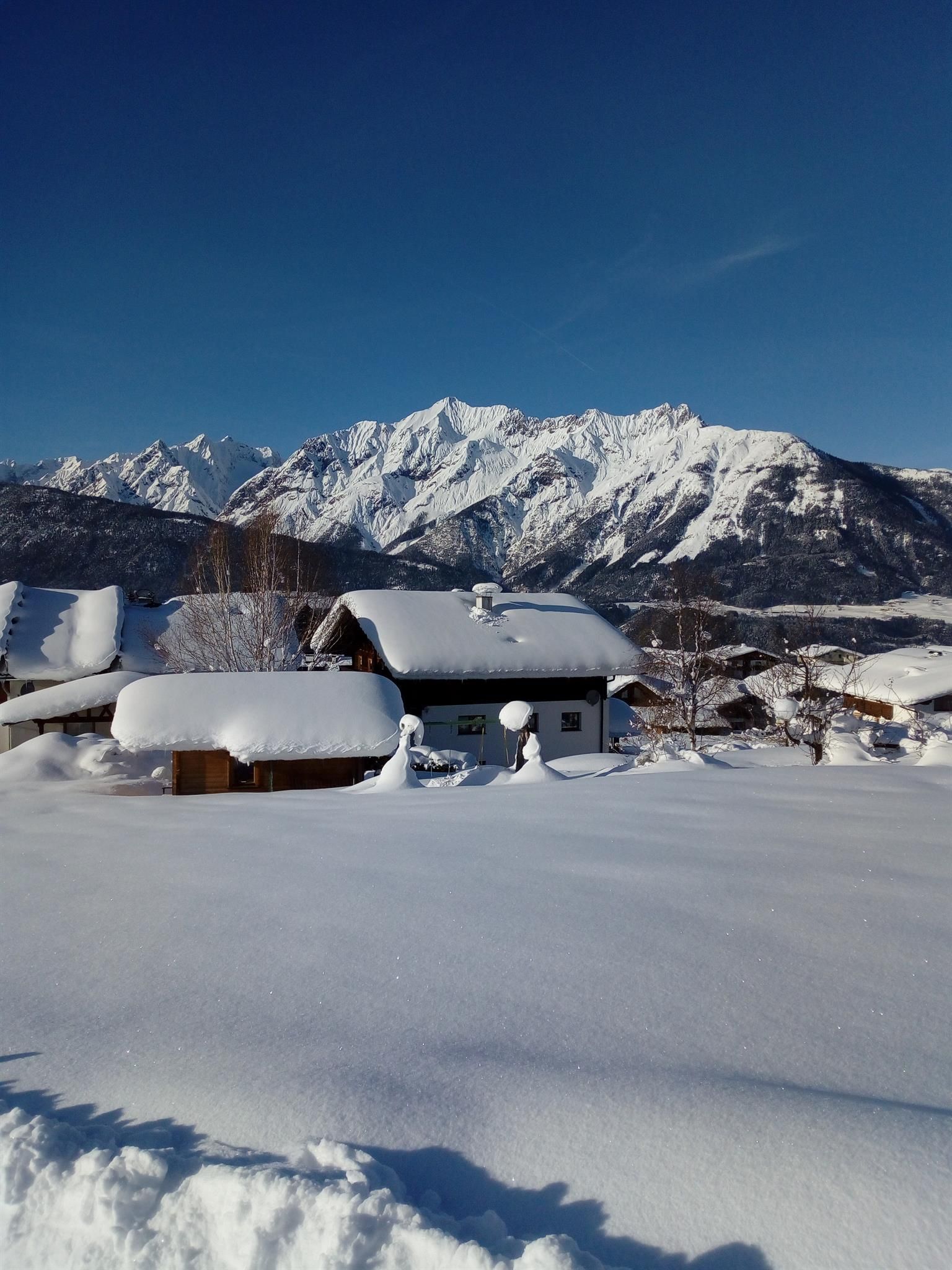 A snowy landscape with a cozy house and majestic mountains in the background. The clear blue sky complements the winter scene.