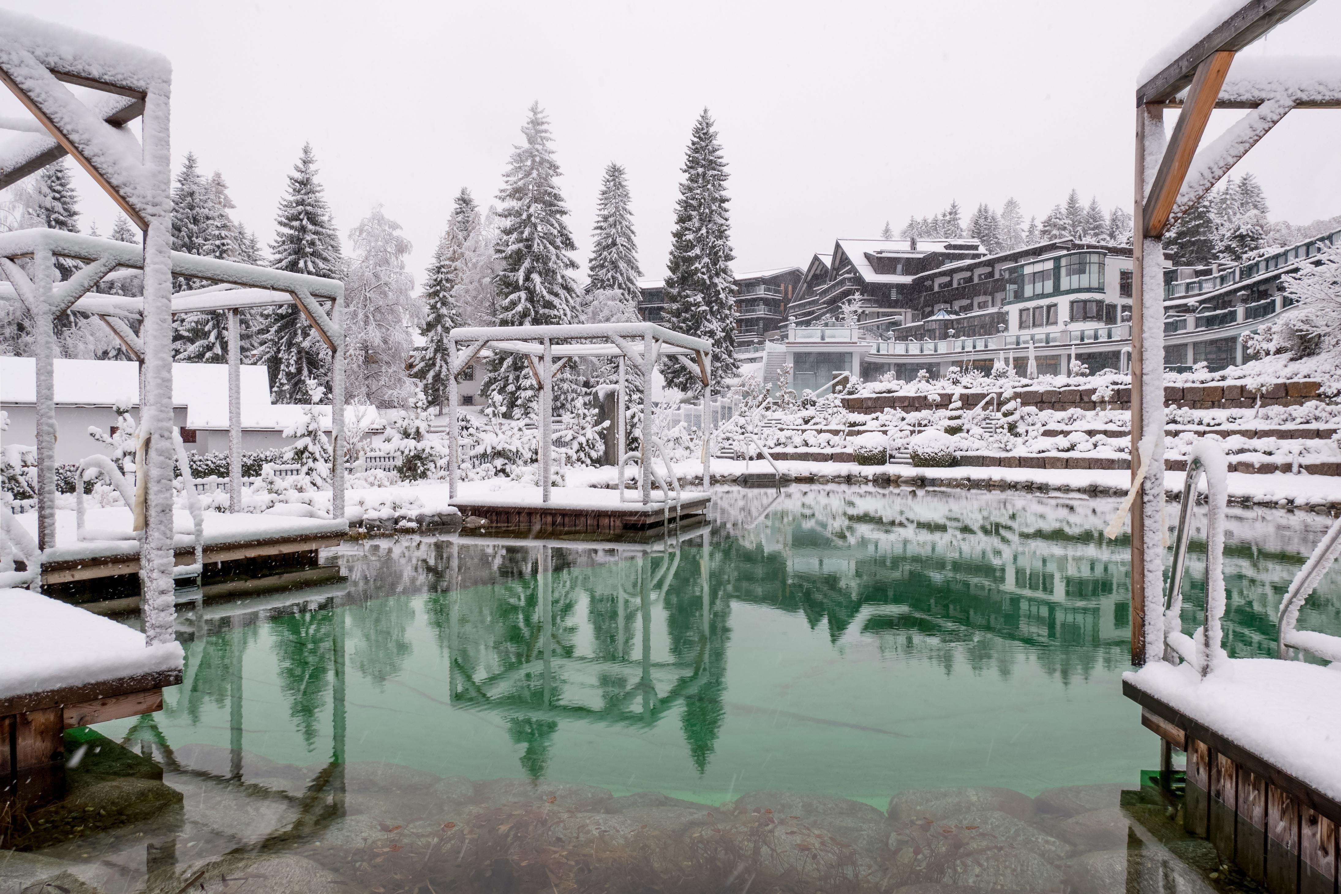 An idyllic winter lake with snow-covered shores and pine trees. In the background, a charming hotel can be seen, surrounded by a snowy landscape.