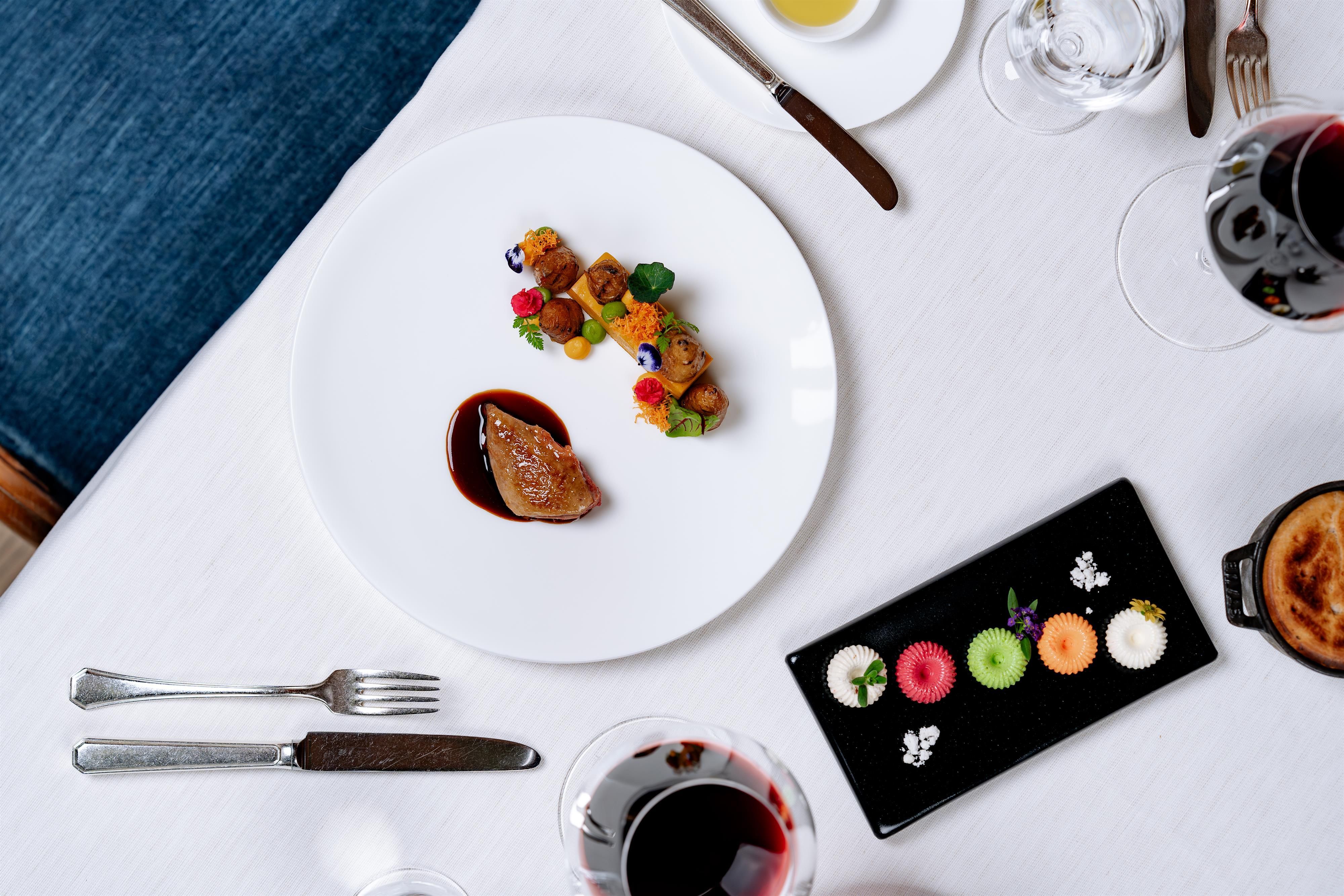 An elegantly arranged plate with meat and colorful vegetables. Next to it, a selection of small desserts on a black plate.