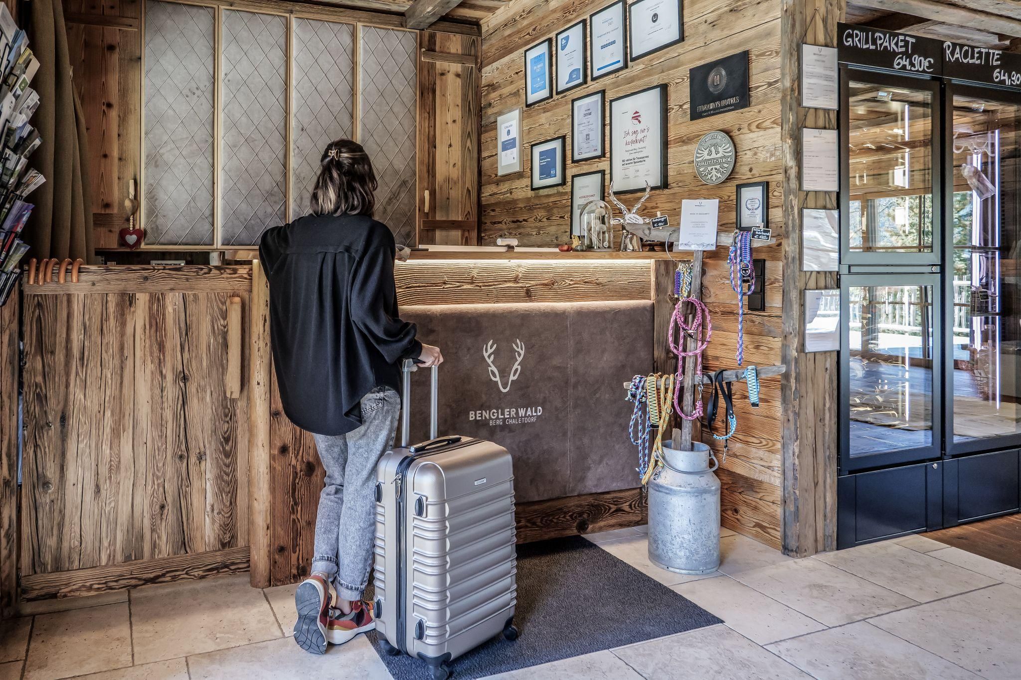 A person stands at the reception of the chalet village with a suitcase. Various awards and posters are hanging on the wall.