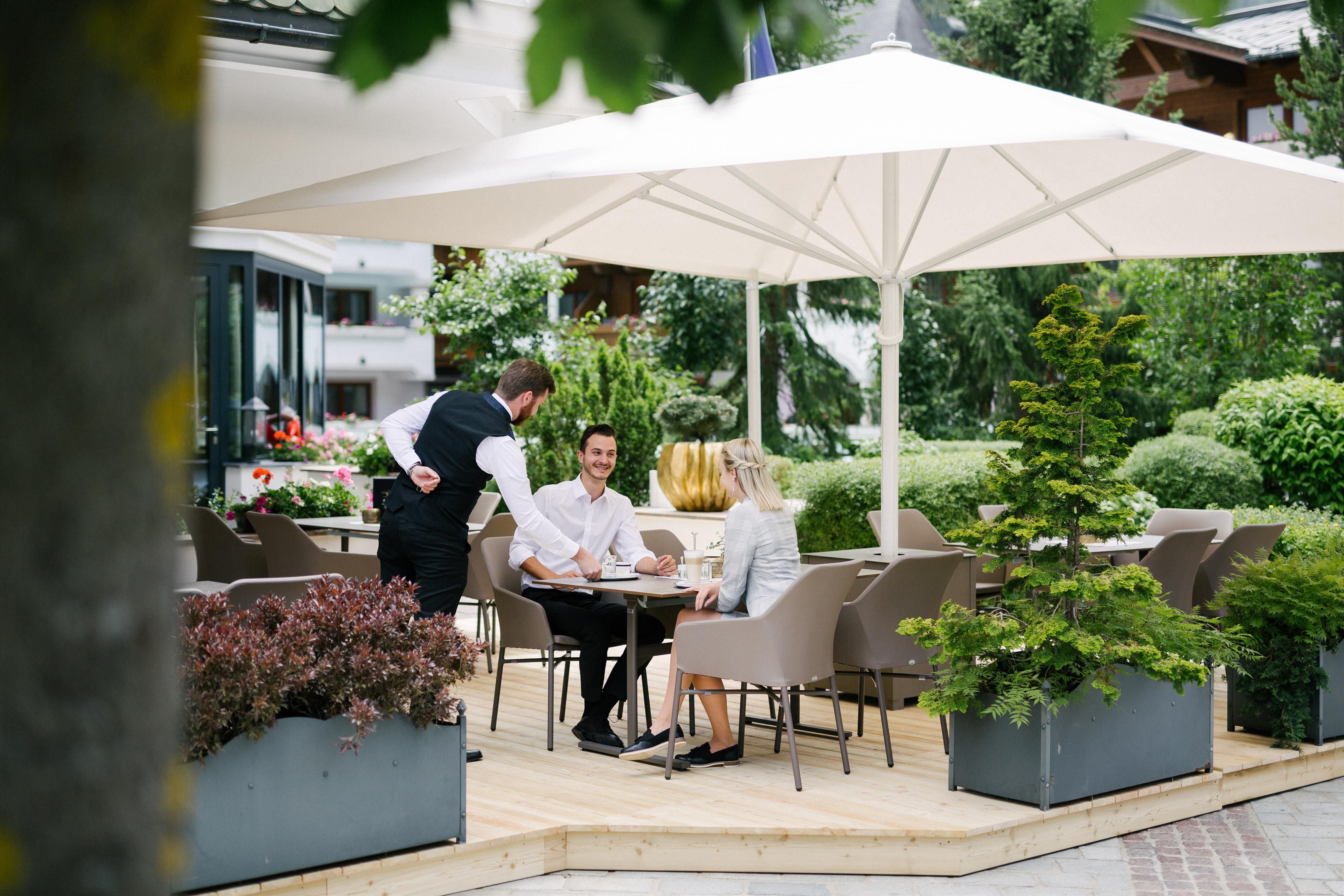 A cozy outdoor area with a table and chairs under a sunshade. A waiter serves two guests in a green, inviting environment.
