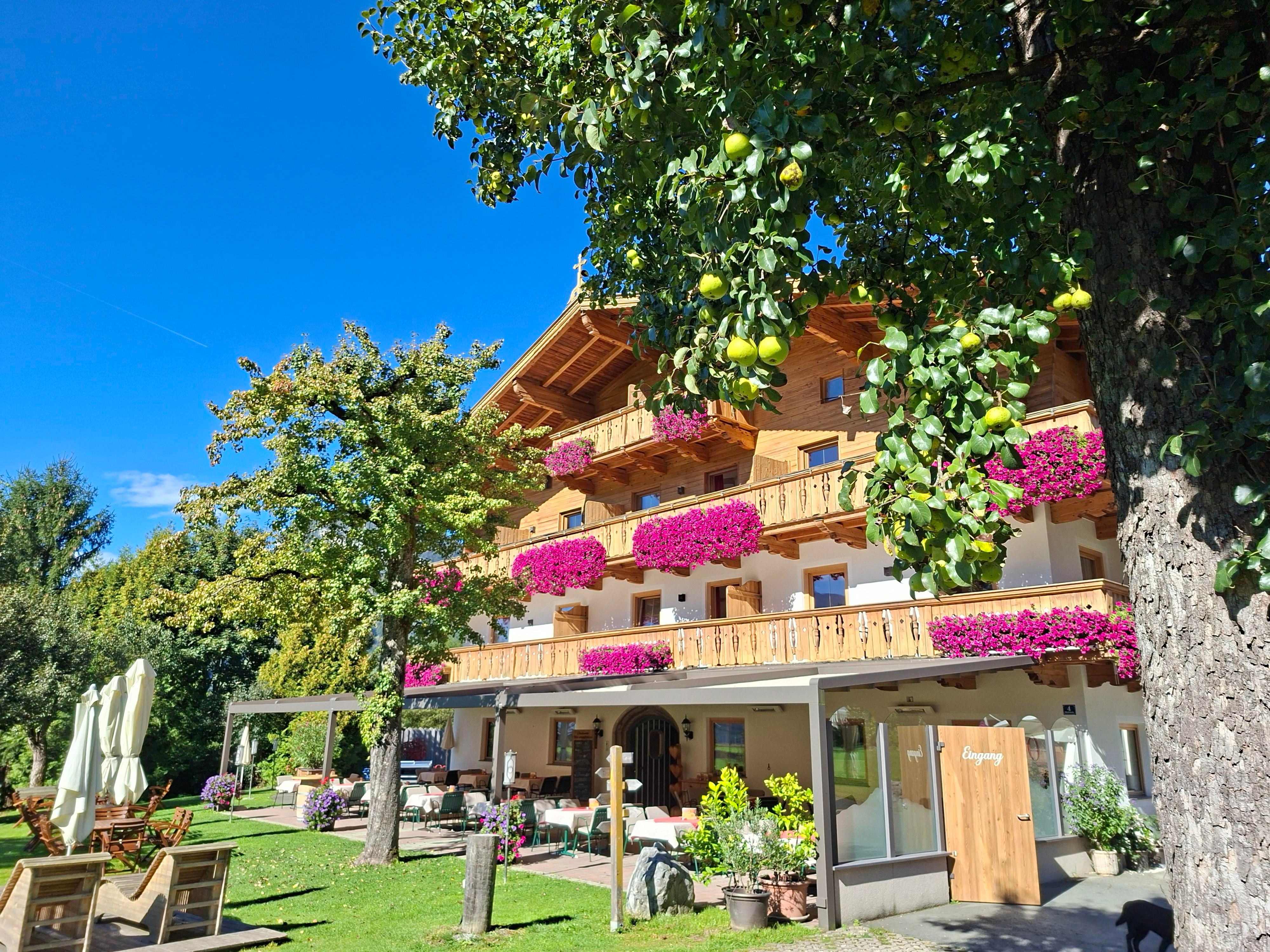 A charming wooden house with balconies full of flowers and a beautiful garden. The sky is blue and the surroundings are green and inviting.