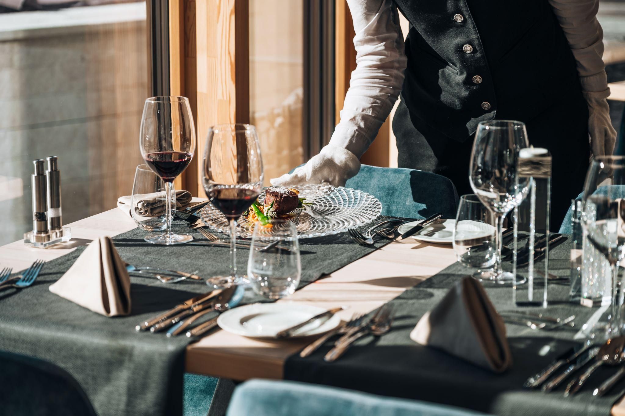 An elegantly set table in a restaurant with wine glasses and fine table decoration. A waiter is serving a dish on a decorative plate.