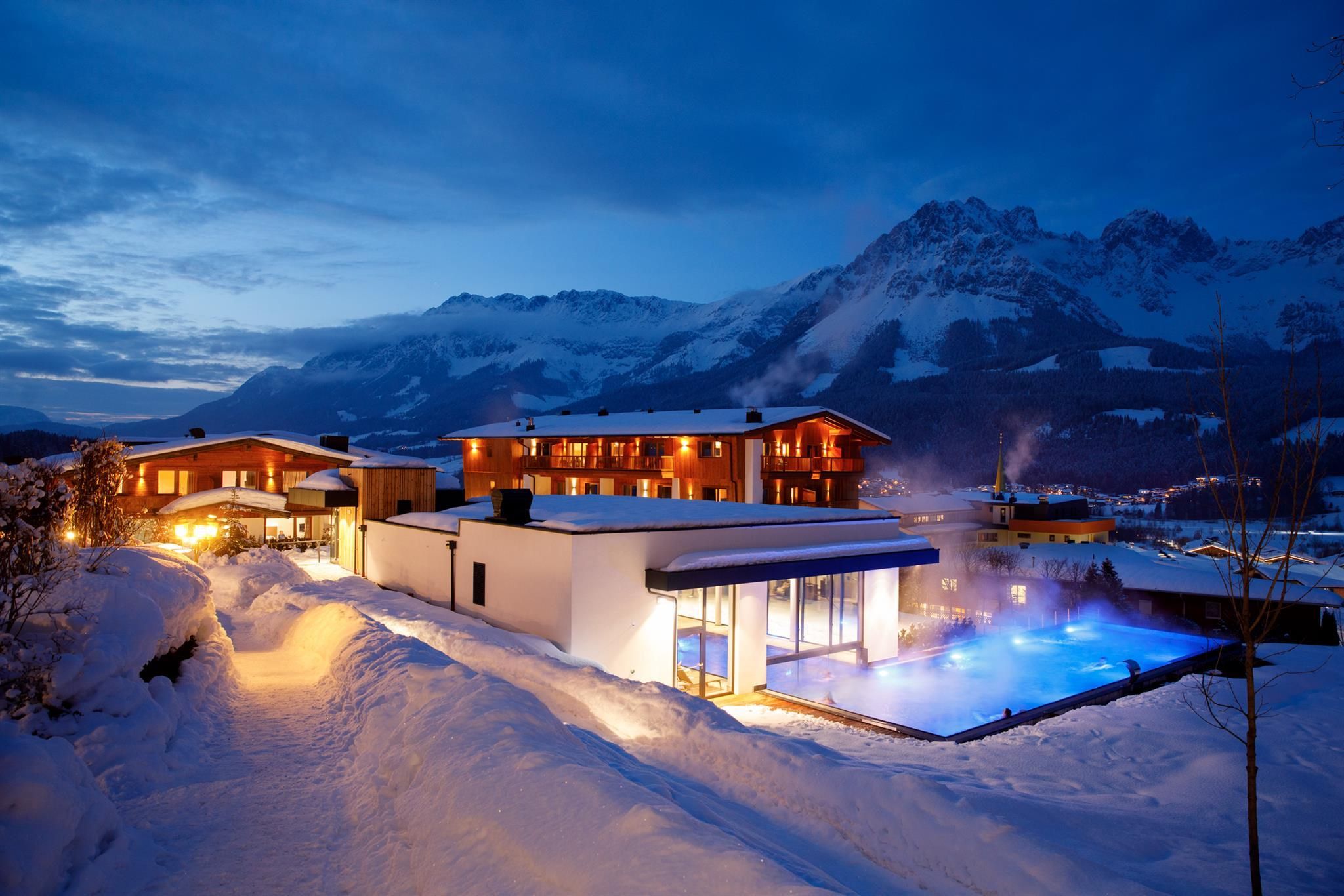 A modern hotel in a snowy mountain landscape at twilight. The outdoor area features a lit pool and snow-covered landscapes.