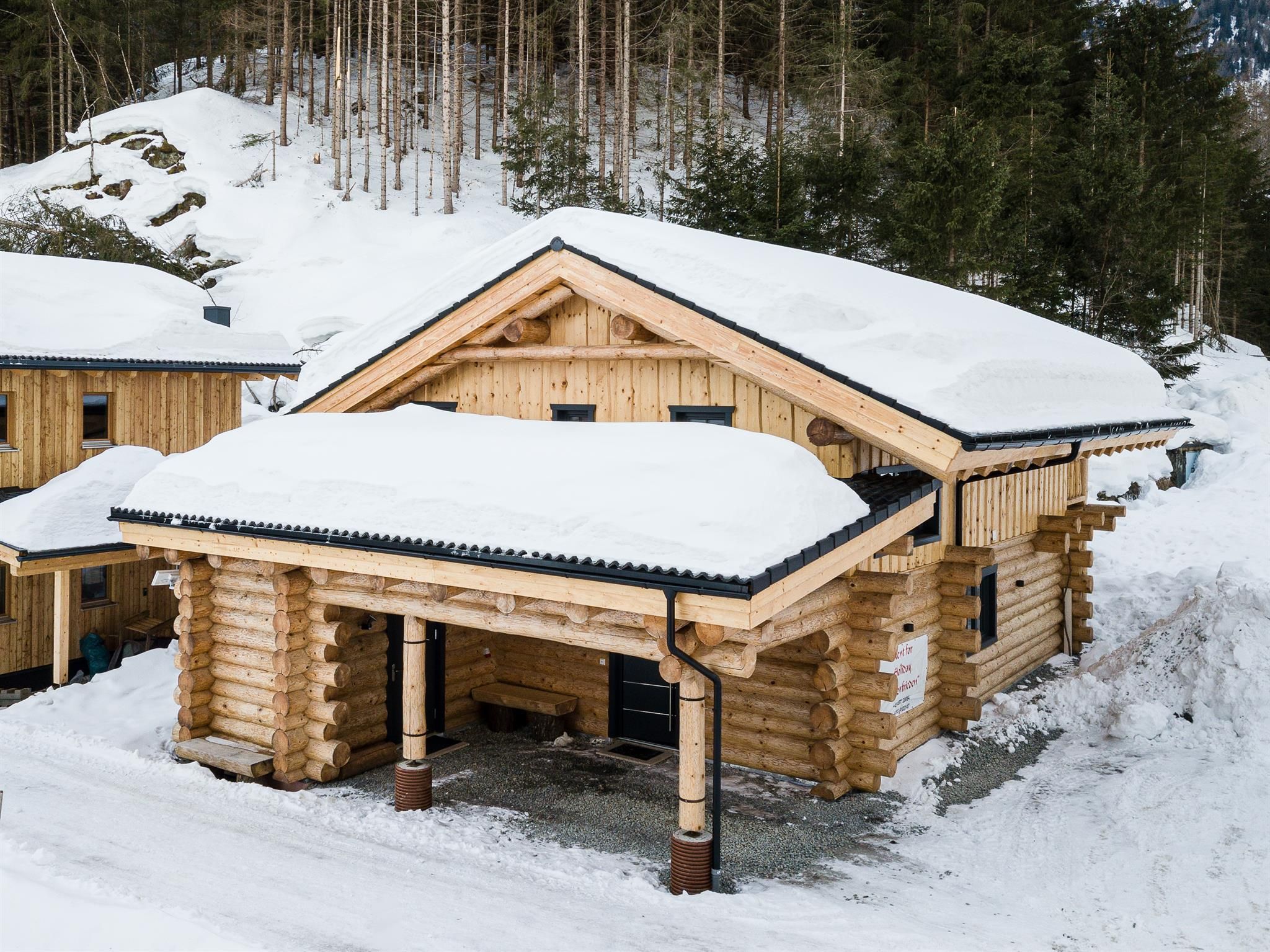 A wooden house with a snow-covered roof stands in a winter landscape. Surrounded by trees, it appears cozy and inviting.