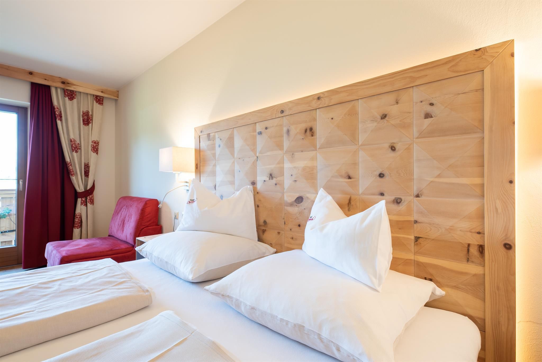 A cozy bedroom with a wooden bed and large, comfortable pillows. On the wall, there is an artistic wooden paneling, and a red armchair is placed next to the window.
