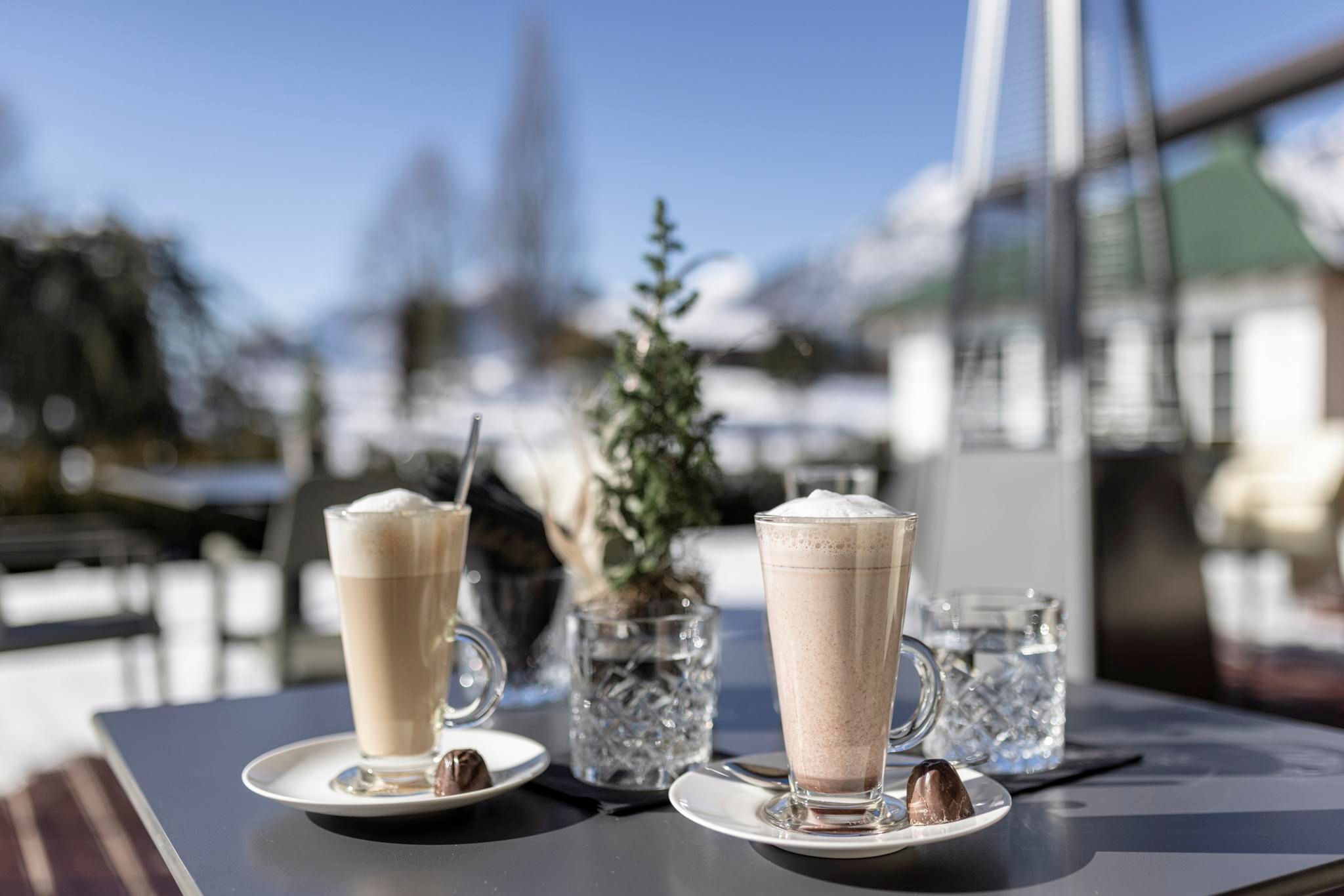 Two glasses of coffee with cream on an outdoor table. In the background, there are trees and snow-capped mountains visible.
