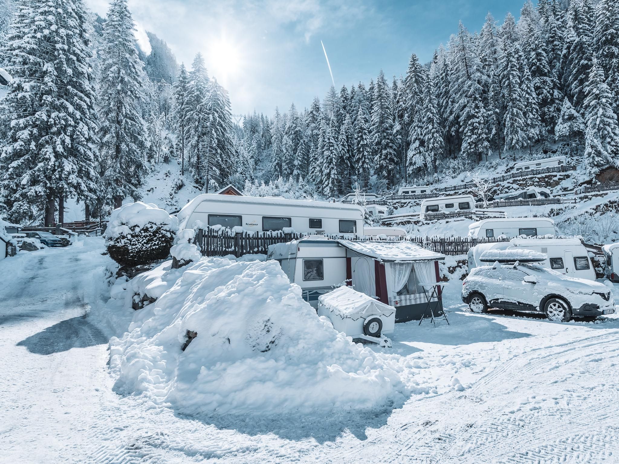 A snowy landscape with cozy caravans and tall, snow-covered trees. The sun is shining, and there is a lot of snow on the ground.