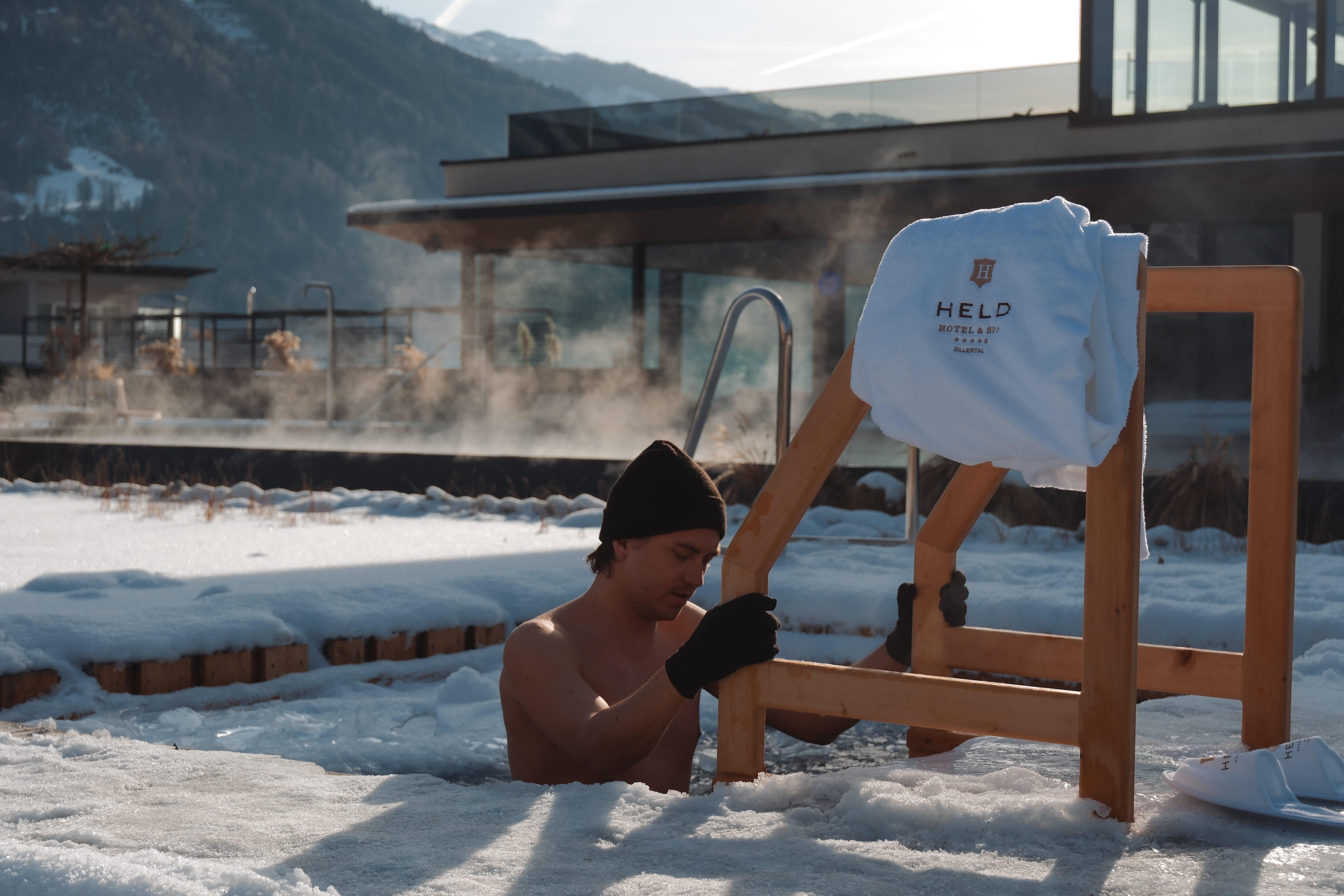 A man is sitting in a warm spring outdoors, surrounded by snow and mountains. Steam clouds are rising and a towel with the inscription "HERO" is lying on a chair.