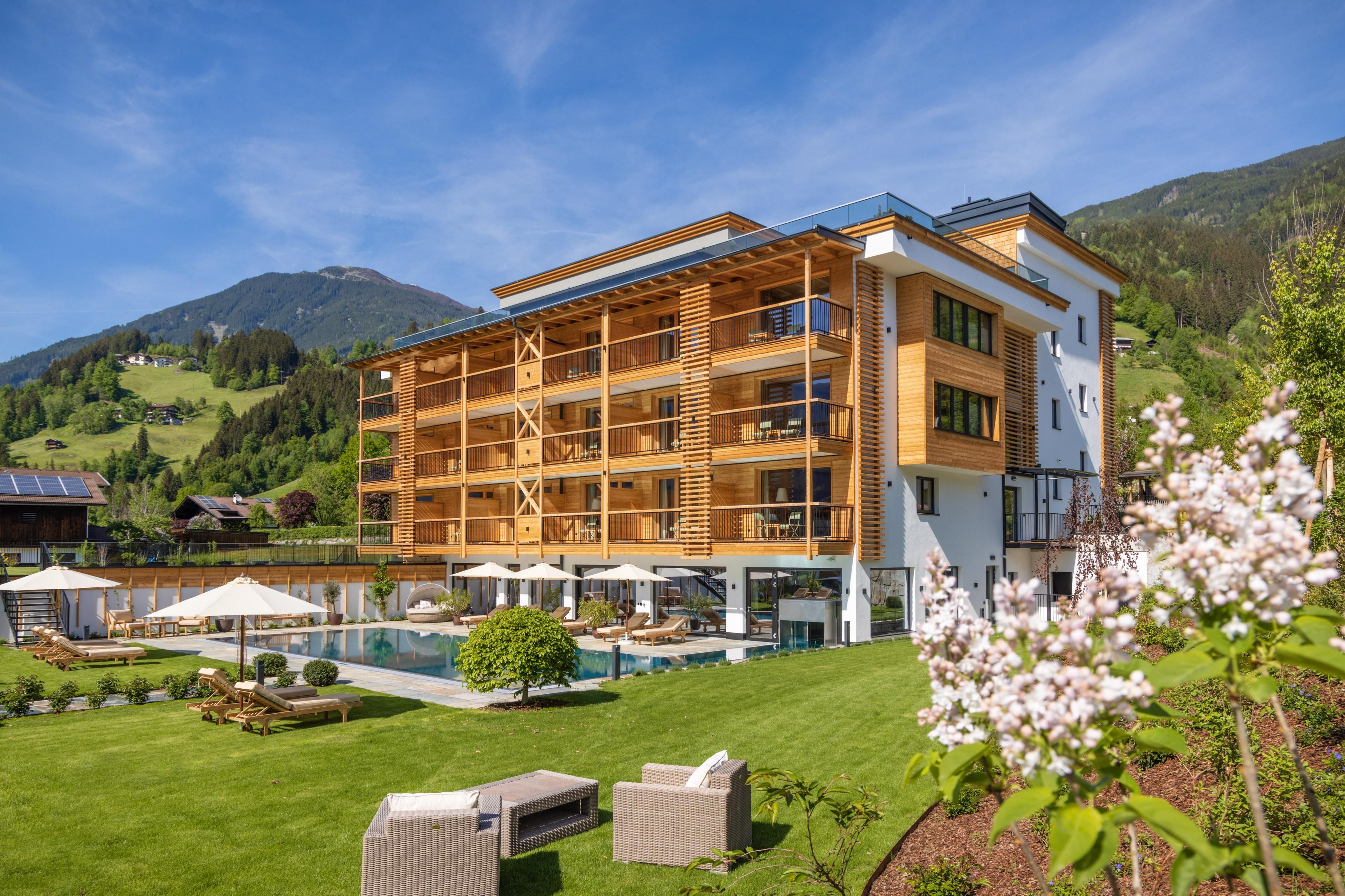 A modern hotel building surrounded by green hills and blooming flowers. The pool area invites you to relax.