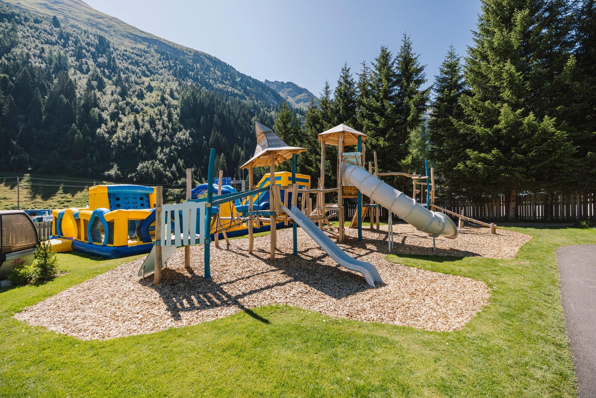 A playground with slides and climbing frames, surrounded by green spaces and trees. In the background, mountains can be seen.