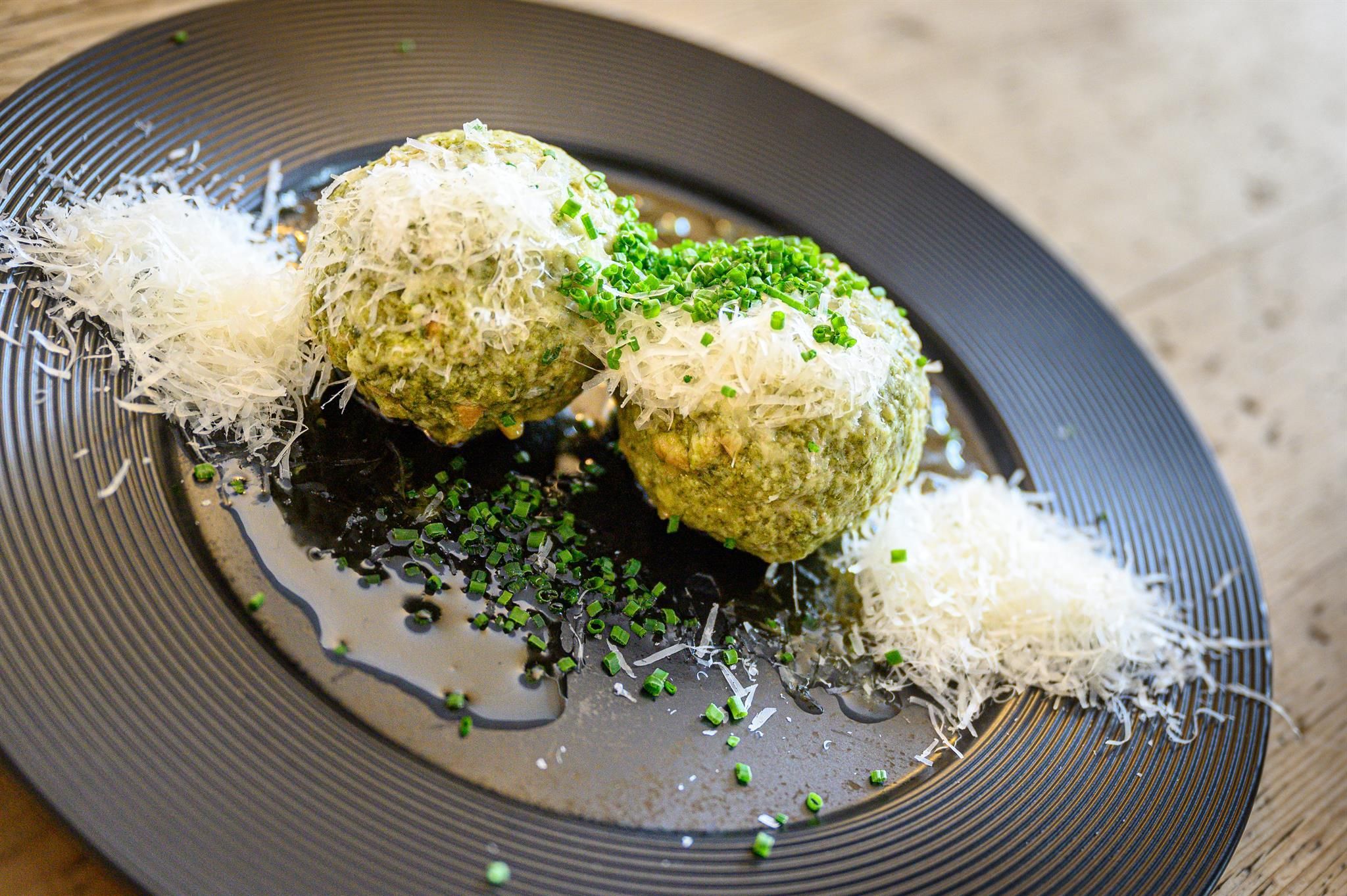 Two green risotto balls on a black plate, garnished with cheese and fresh chives. The plate is artistically arranged and looks appetizing.