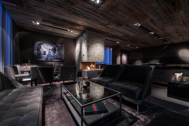 A stylish living room with dark furniture and a modern fireplace. The space has large windows and a cozy atmosphere.