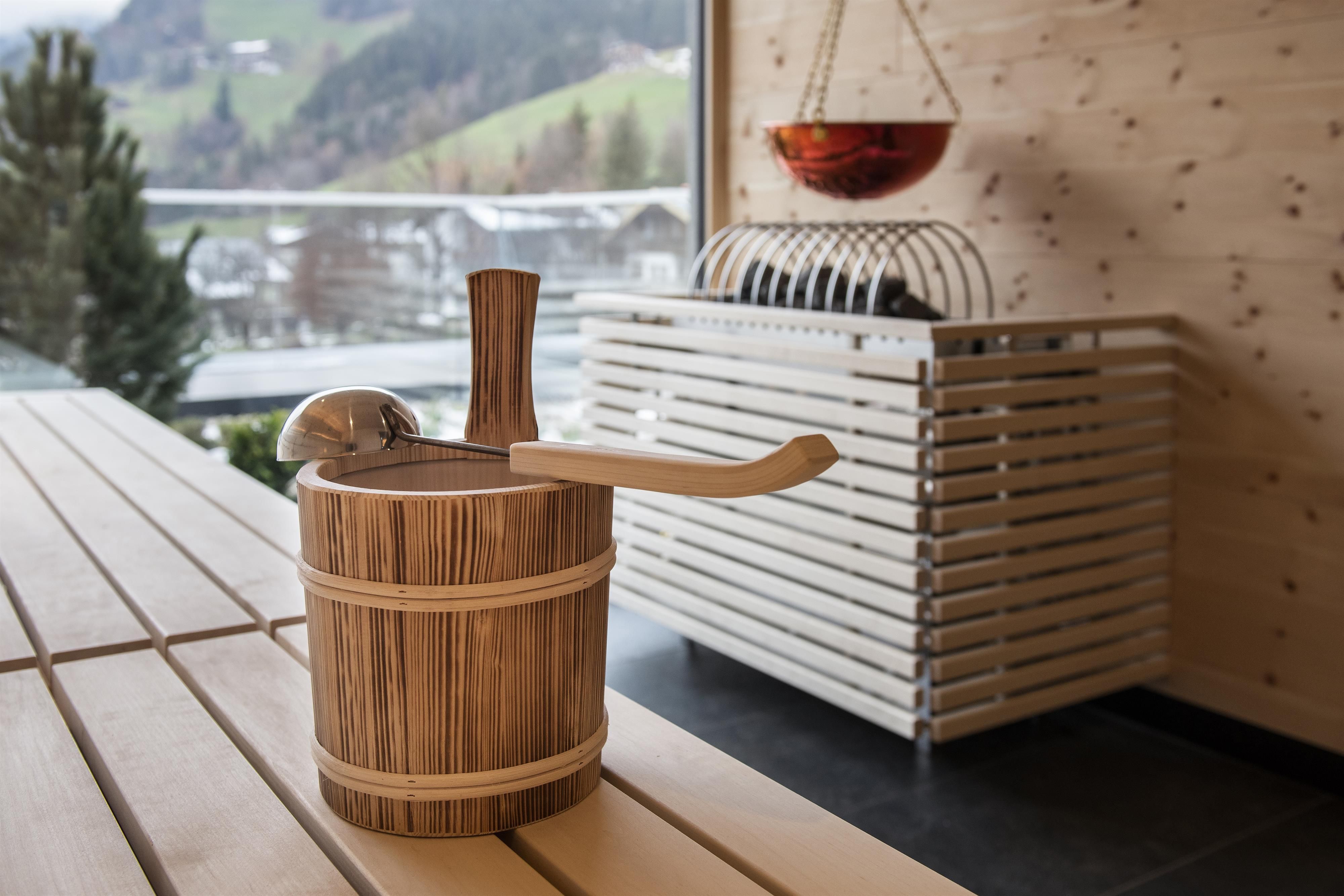 A wooden bucket is on a table in a modern room. In the background, wooden walls and a sauna are visible.