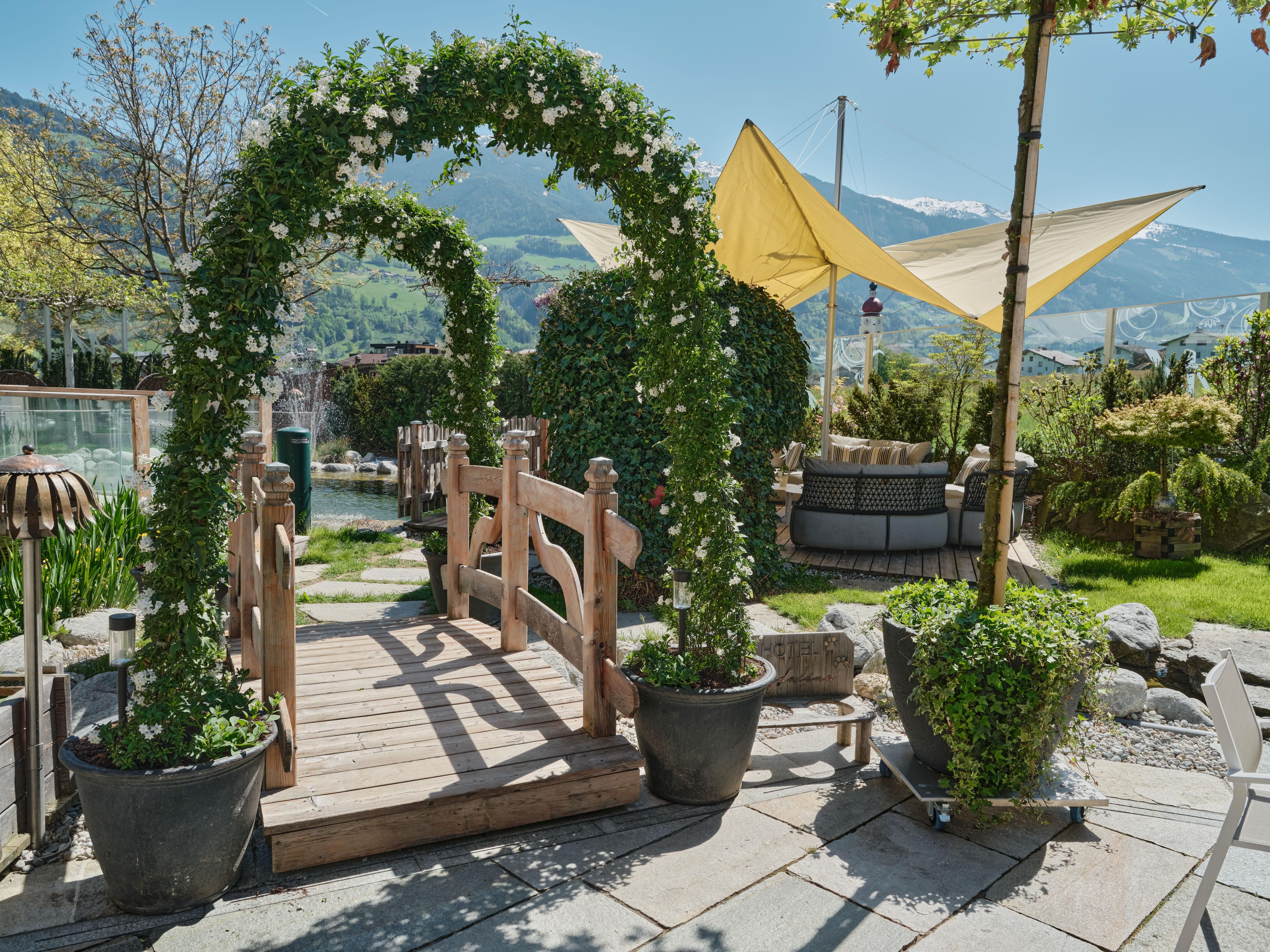 A beautiful wooden bridge leads through an arch decorated with flowers. In the background, green plants and a sunny sky can be seen.