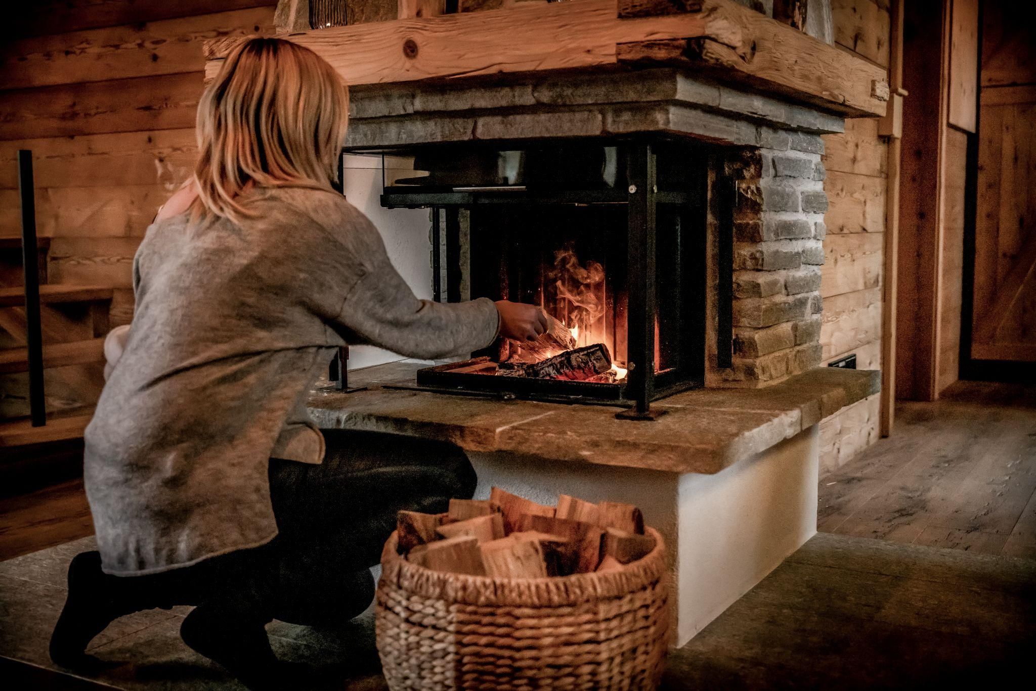 A person is sitting in front of a fireplace and feeding the fire with wood. In the background, wooden paneling and a basket with logs are visible.