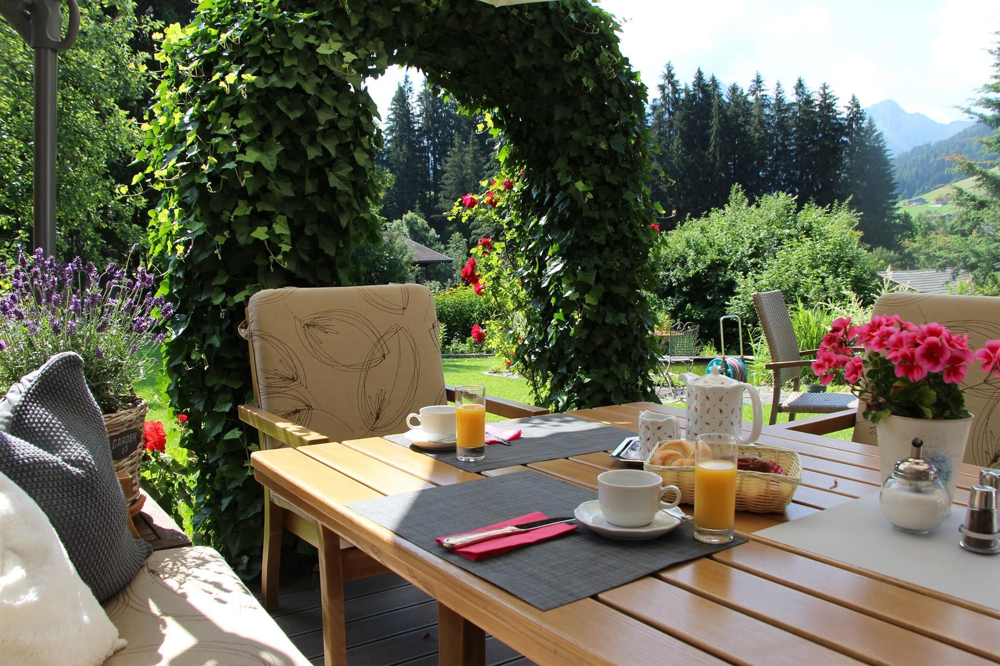 A beautiful outdoor breakfast table, surrounded by blooming plants and green trees. The landscape in the background features an impressive mountain backdrop.